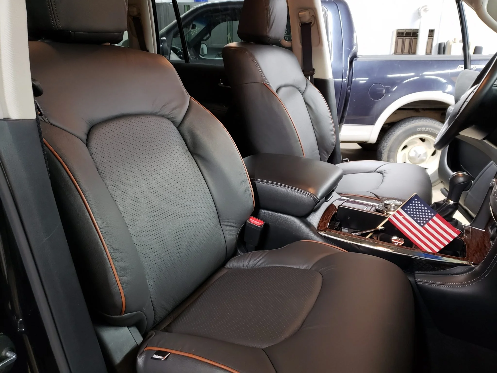 The interior of a vehicle showing black leather seats with orange piping, a dashboard, and an American flag decoration on the center console. There is a view of another vehicle through the front windshield.