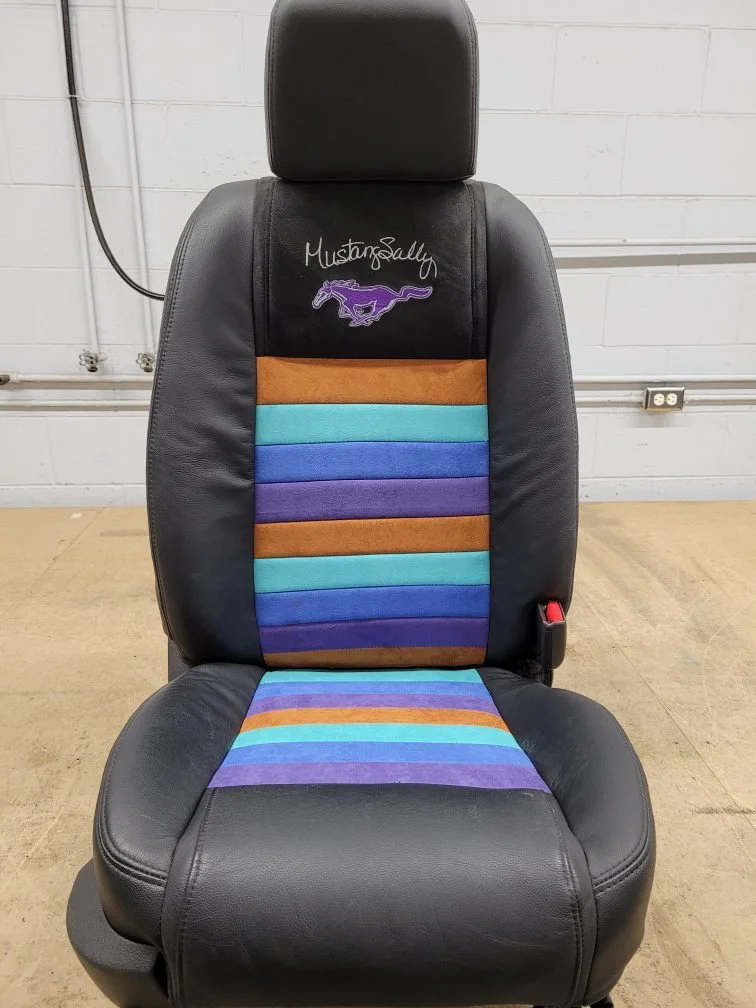 A black car seat with embroidered Mustang logo and name on the headrest, featuring colorful striped fabric with orange, teal, purple, and blue patterns on the seat back and bottom cushion.