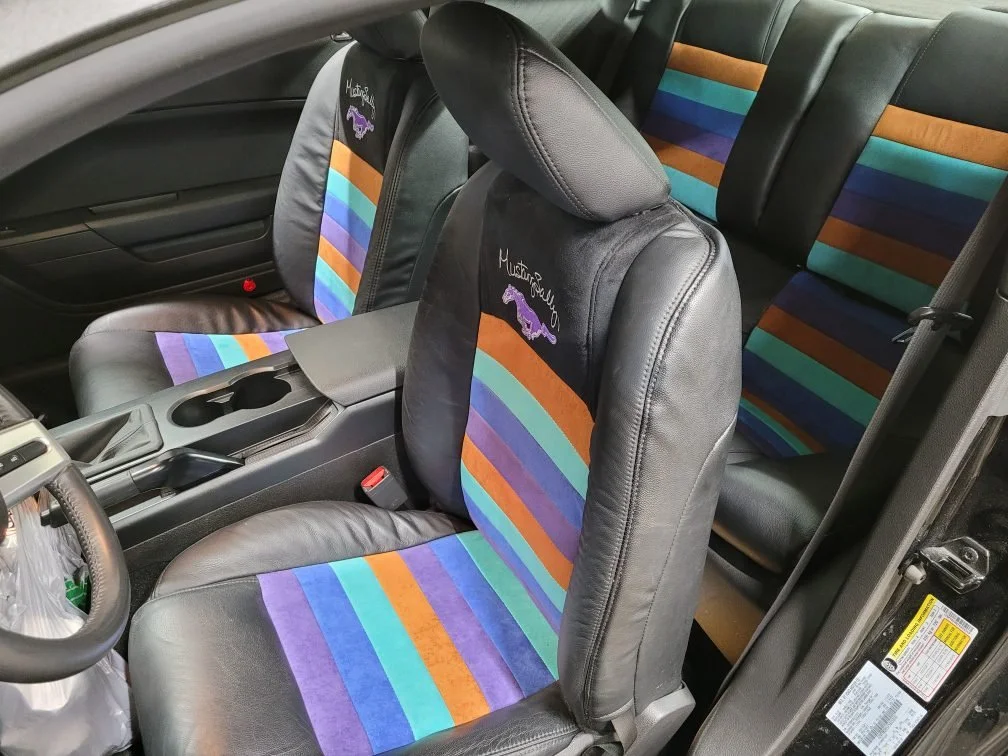 The interior of a vehicle, showing black leather seats with colorful striped suede in seats. The seats have purple, blue, green, orange, and teal stripes. The seat covers have personalized embroidered text and a purple Mustang logo.