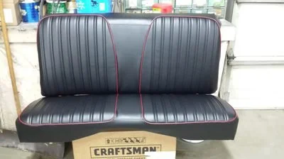 A black vintage car bench seat with red piping in a workshop setting.