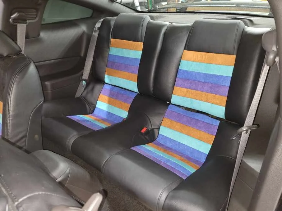 The back seats of a black car with colorful fabric inserts, including orange, purple, and teal stripes, on the seat and backrest.