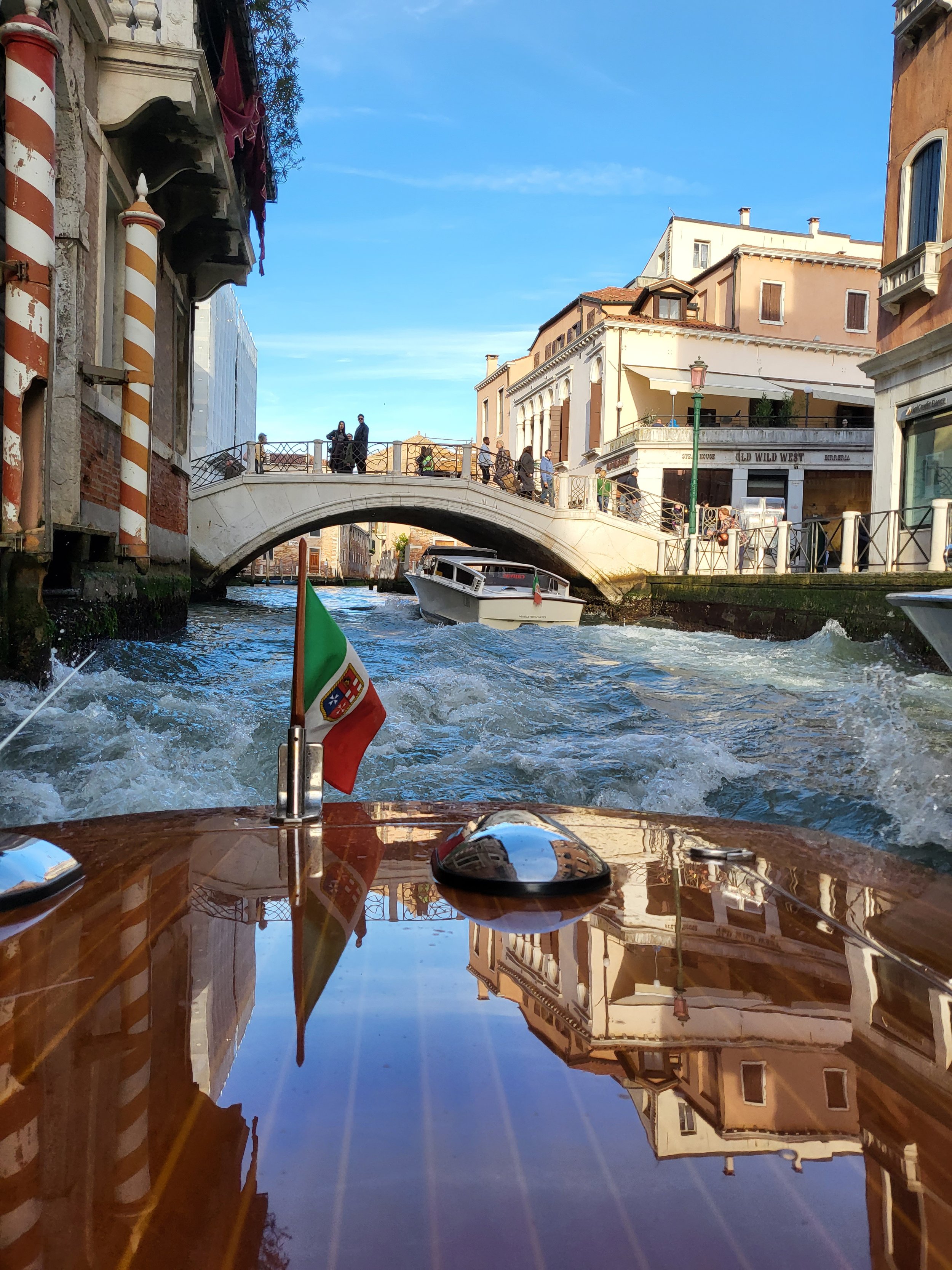 View from a boat on a canal showing a small white boat in the water, a stone bridge with pedestrians, and buildings with colorful facades and windows reflecting on the water.