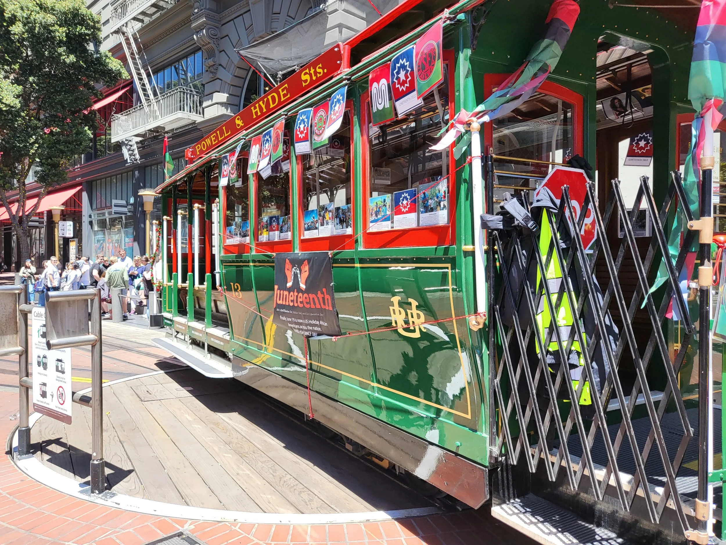 A decorated streetcar or trolley with red and green colors, displays and banners celebrating Juneteenth, with some crowds of people nearby on a city street.