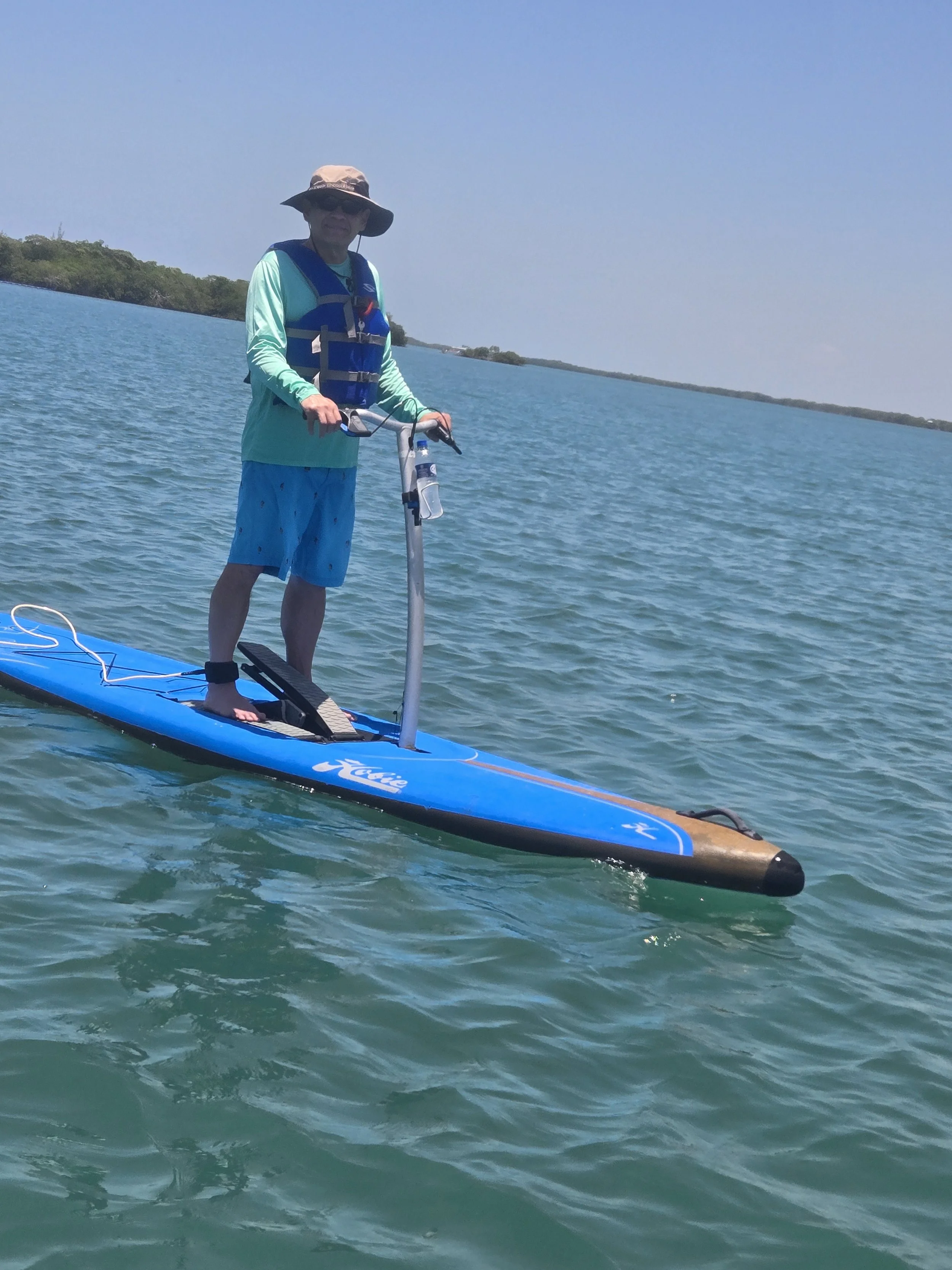 A man paddleboarding on a calm body of water with a distant shoreline and trees in the background, wearing a sun hat, sunglasses, a life jacket, a long-sleeve shirt, and shorts.