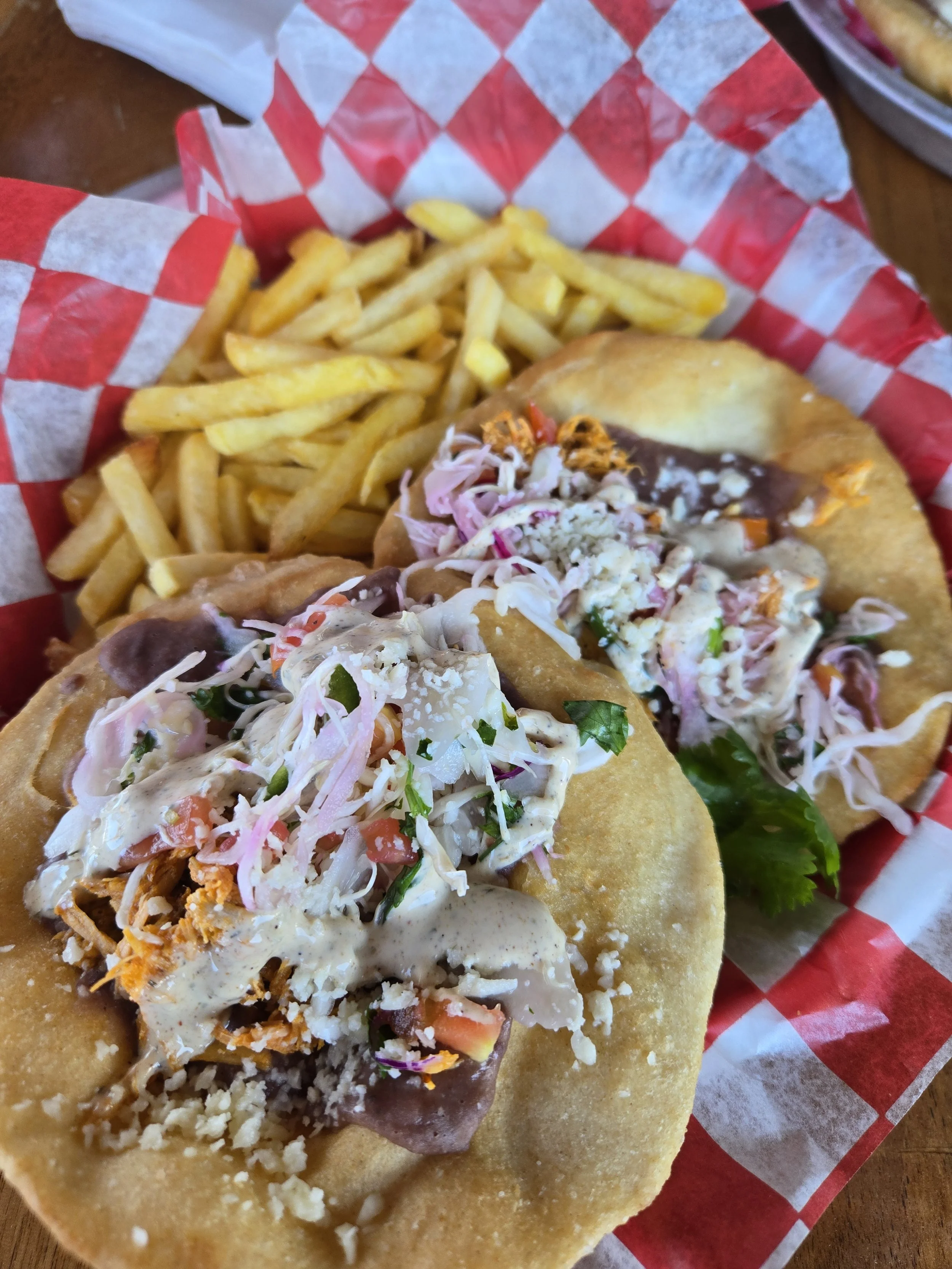 Soft tacos on a round plate with toppings like shredded lettuce, tomatoes, cheese, and sauces, accompanied by a side of French fries on checkered red and white paper