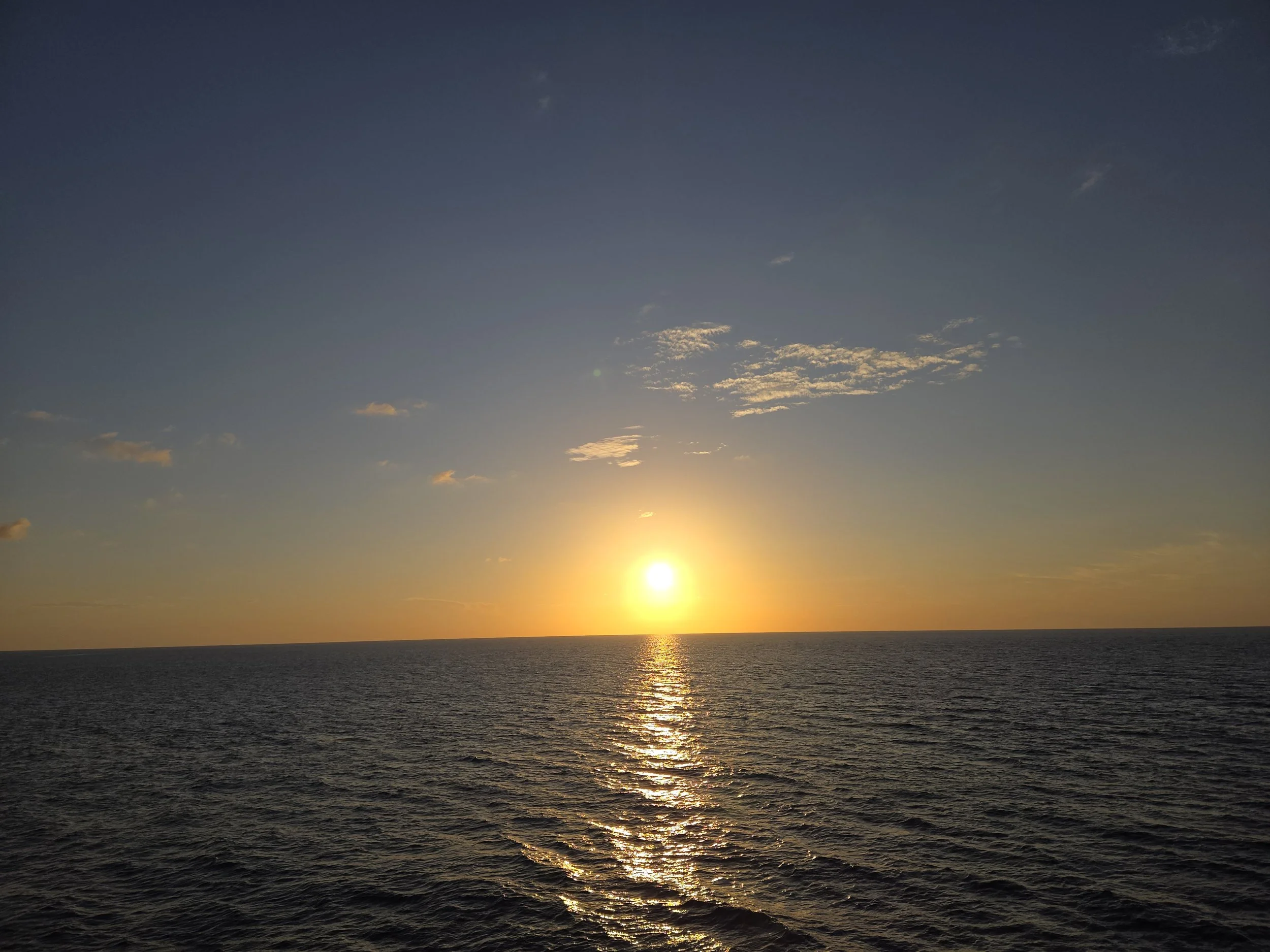 Sunset over the ocean with calm waves and a few scattered clouds in the sky.