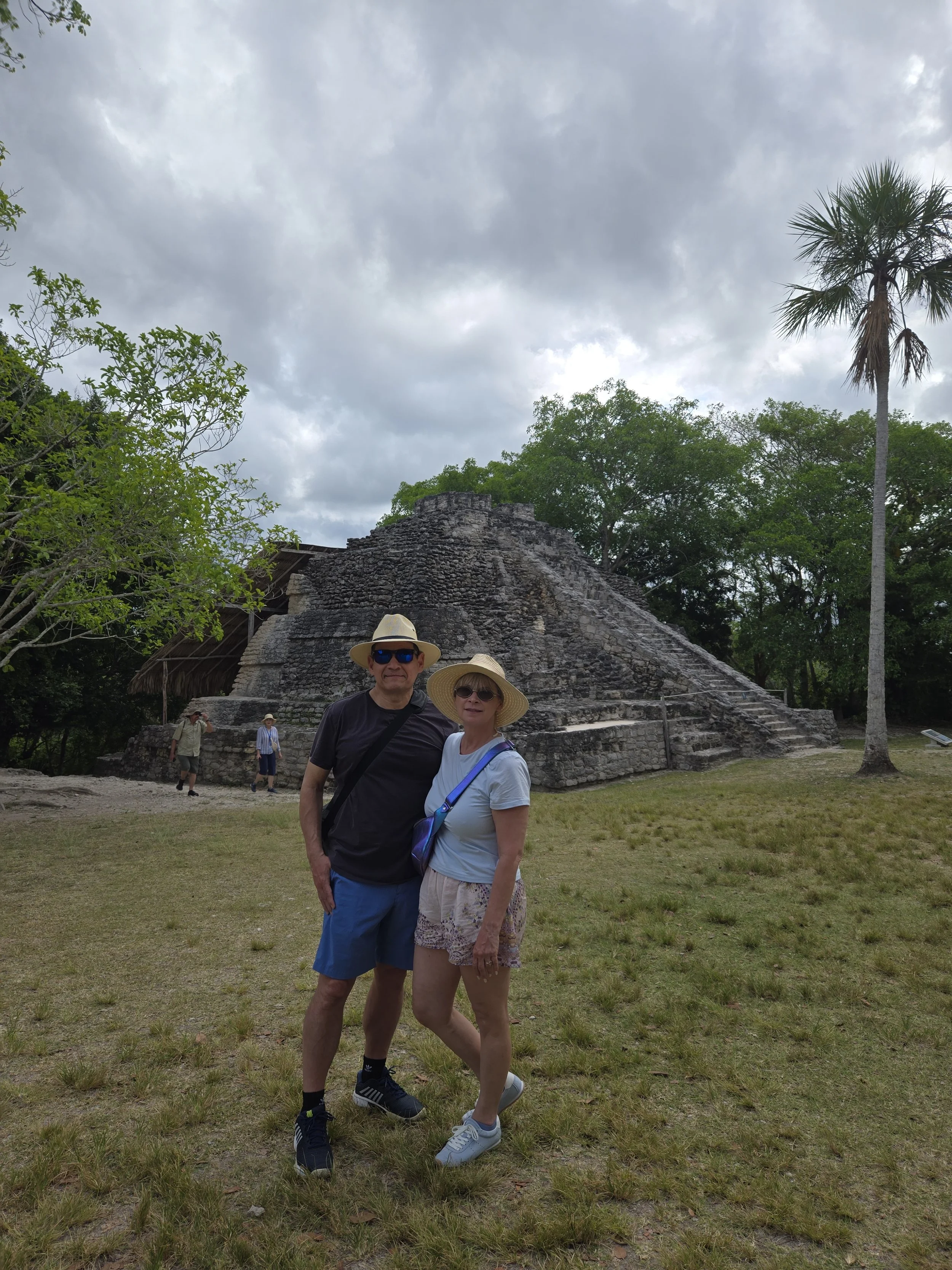 A man and woman standing together outdoors in front of an ancient stone pyramid, wearing hats and sunglasses, with trees and a cloudy sky in the background.