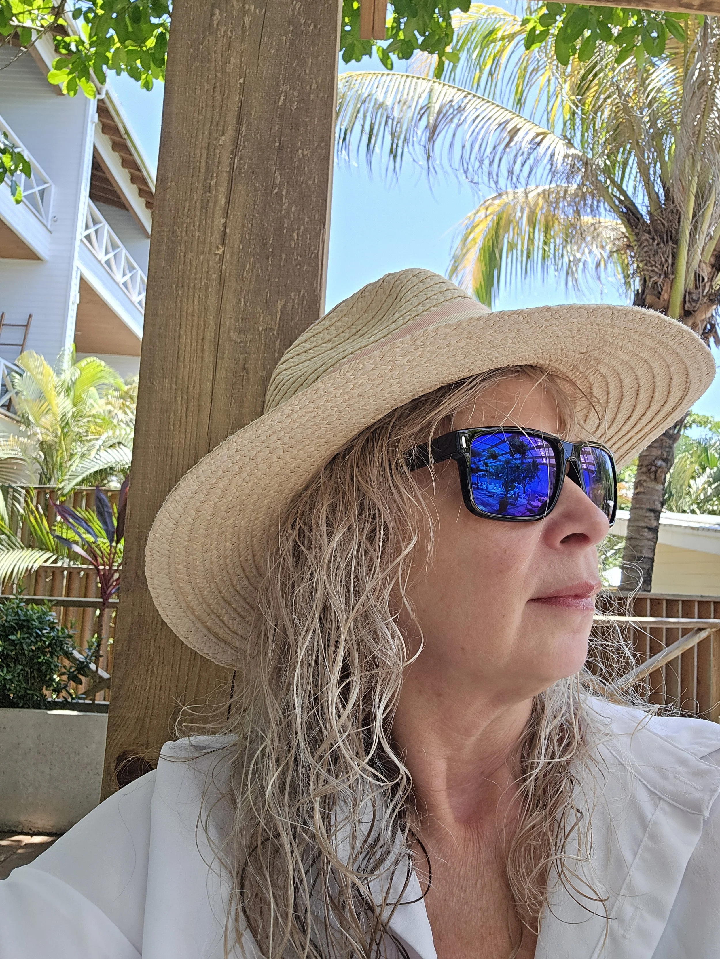 A woman with curly blonde hair wearing a wide-brimmed straw hat and blue reflective sunglasses. She is outdoors with trees, including palm trees, and a building with balconies in the background. Sunny weather.