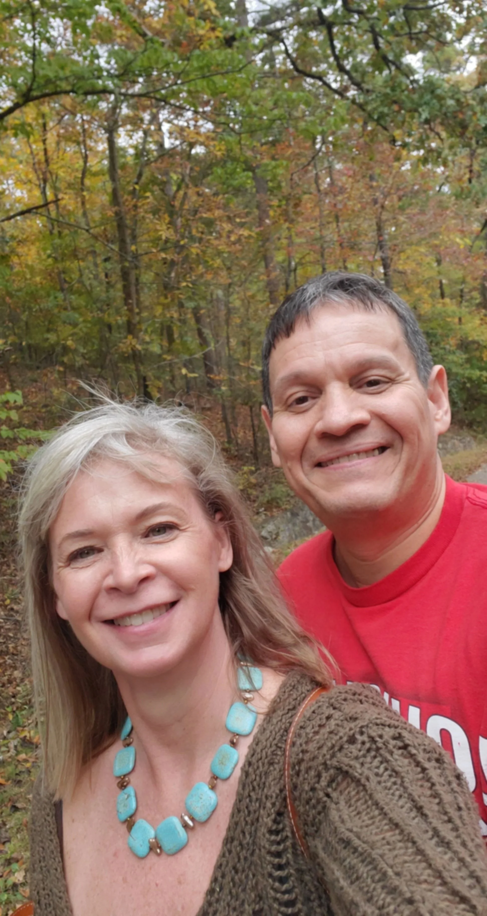 A smiling woman with blonde hair and a turquoise necklace, and a smiling man with short dark hair, wearing a red shirt, taking a selfie outdoors in front of trees with fall foliage.