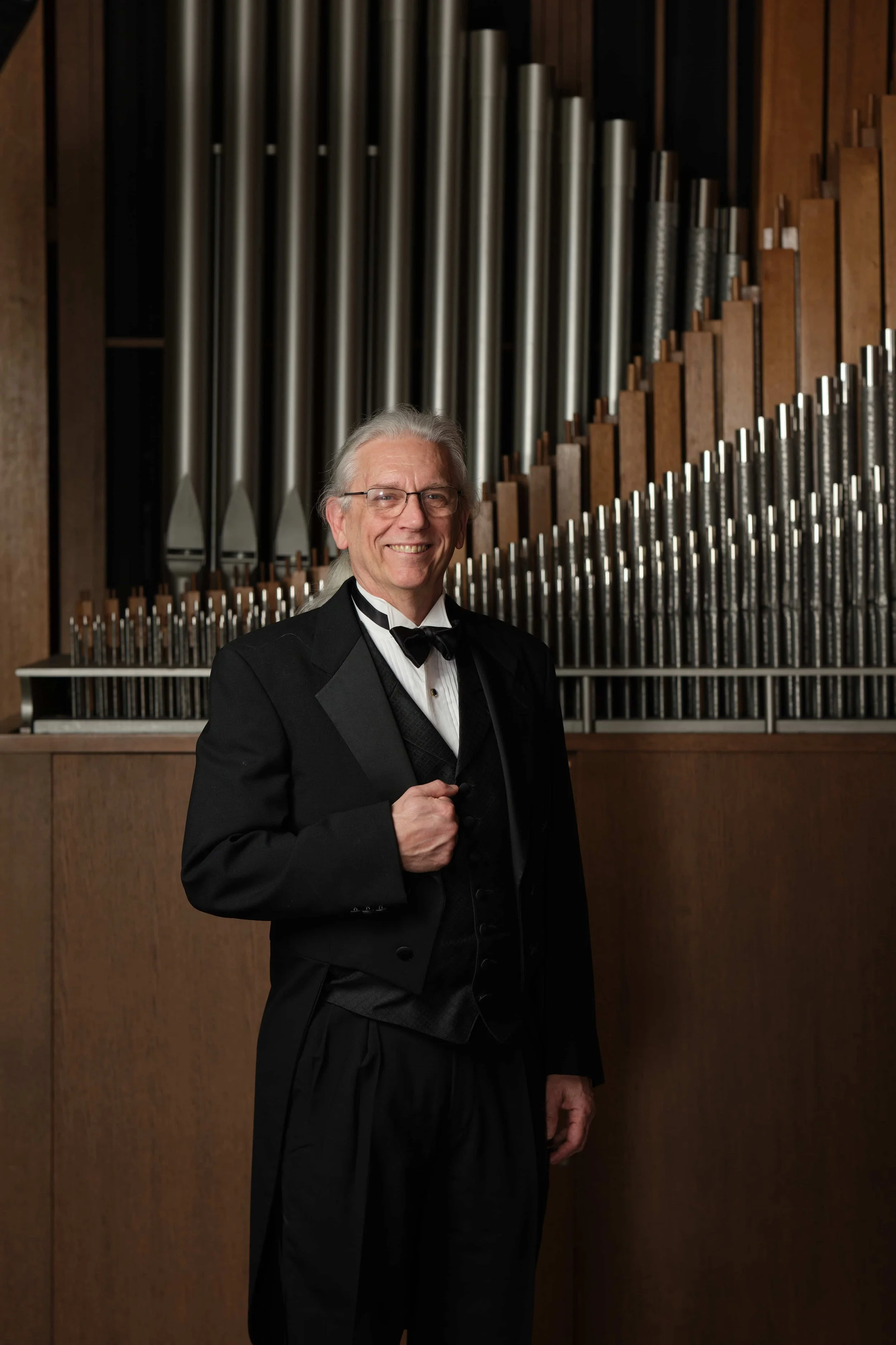 Director of Good Company, Steven Hoifeldt, with gray hair and glasses dressed in a formal tuxedo standing in front of a large pipe organ.