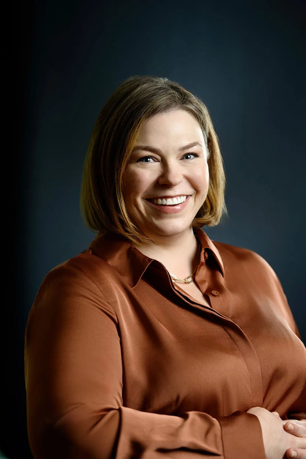 Portrait Julie Musilek, Board President for Good Company, with shoulder-length brown hair, smiling and wearing a copper-colored blouse against a dark background.