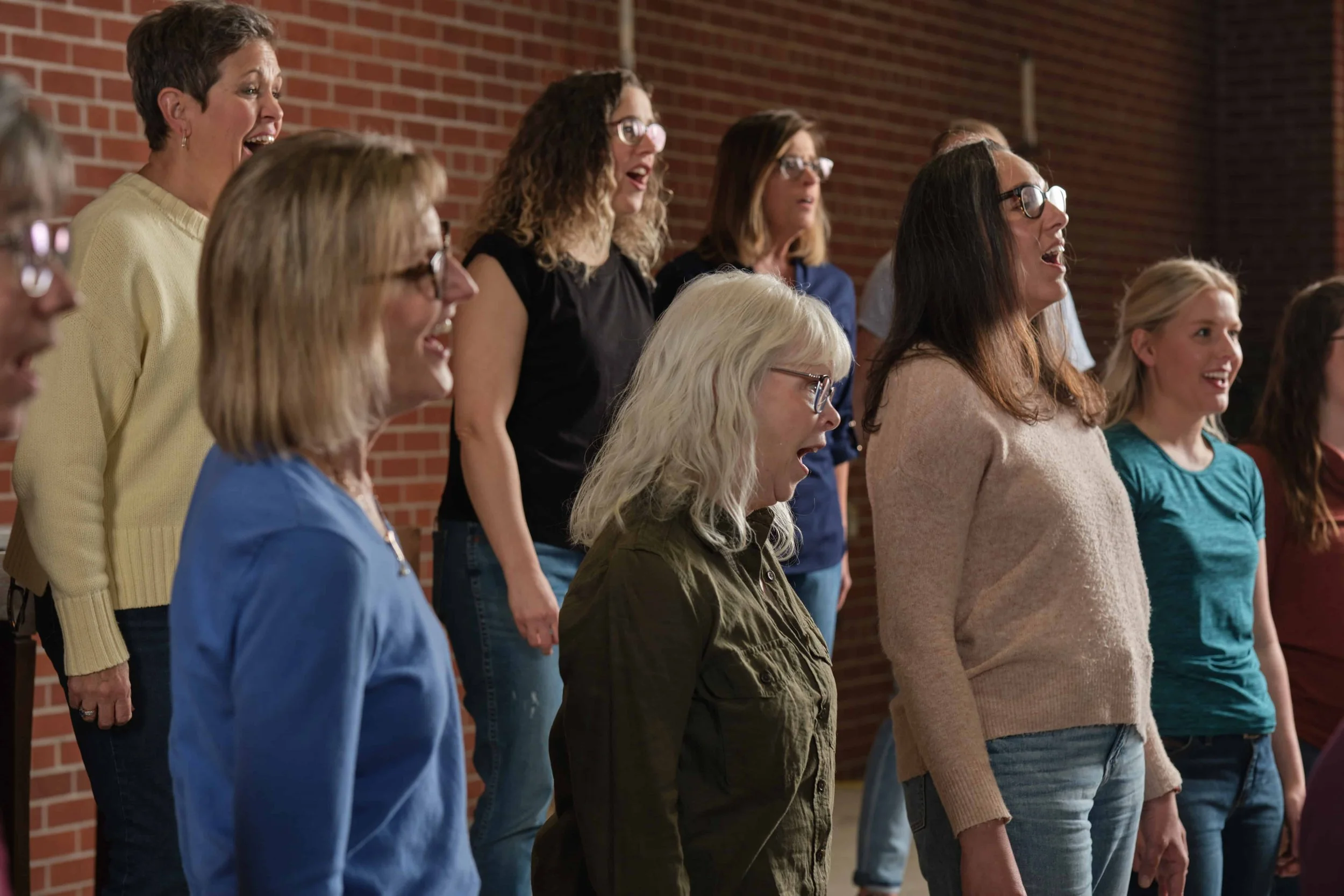 A group of women from Good Comapany, Ames Women's Choir singing together indoors, standing in front of a brick wall.