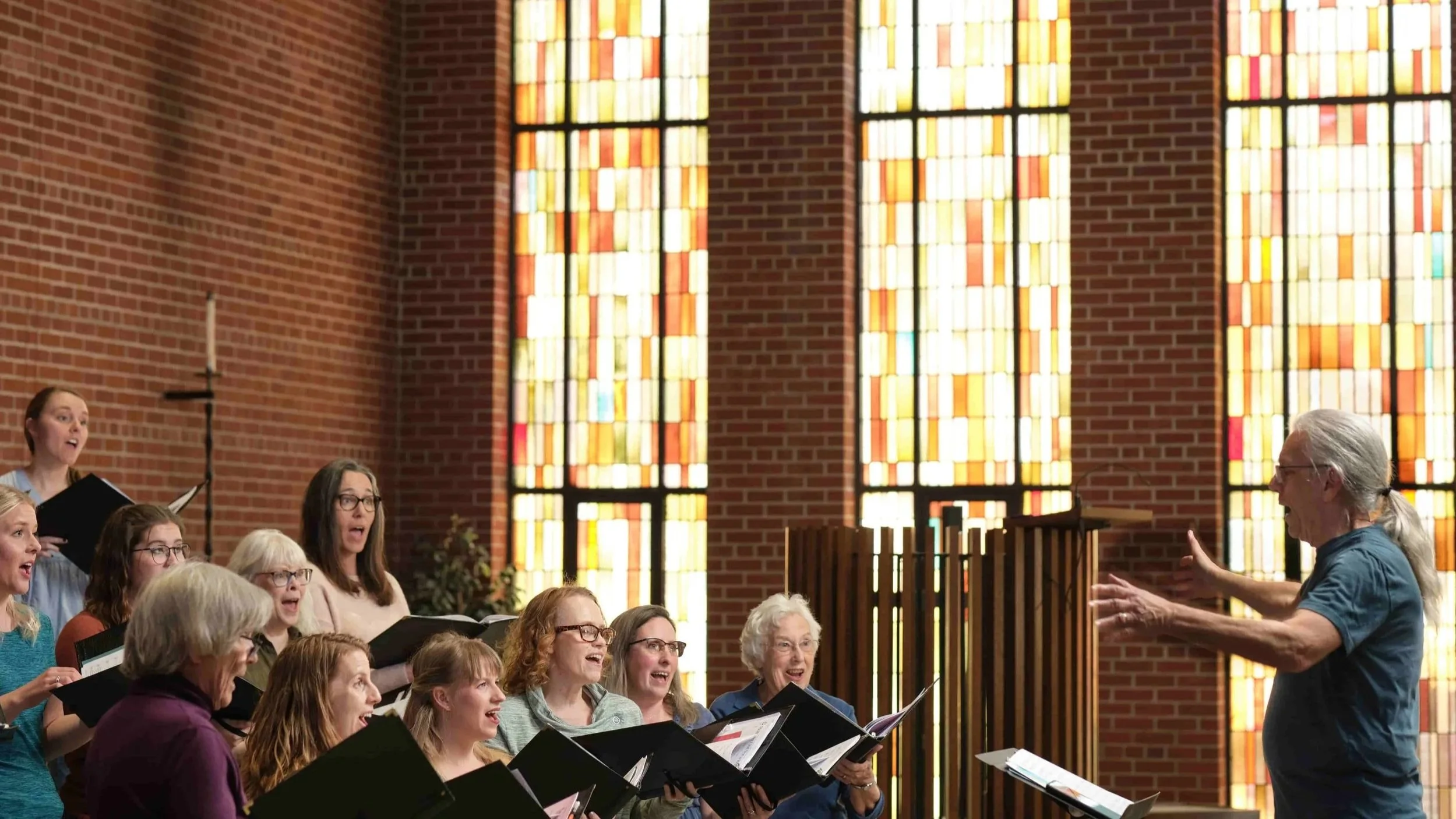 Members of Good Company, Ames Women's Choir singing in a church with stained glass windows with director Steven Hoifeldt leading them.