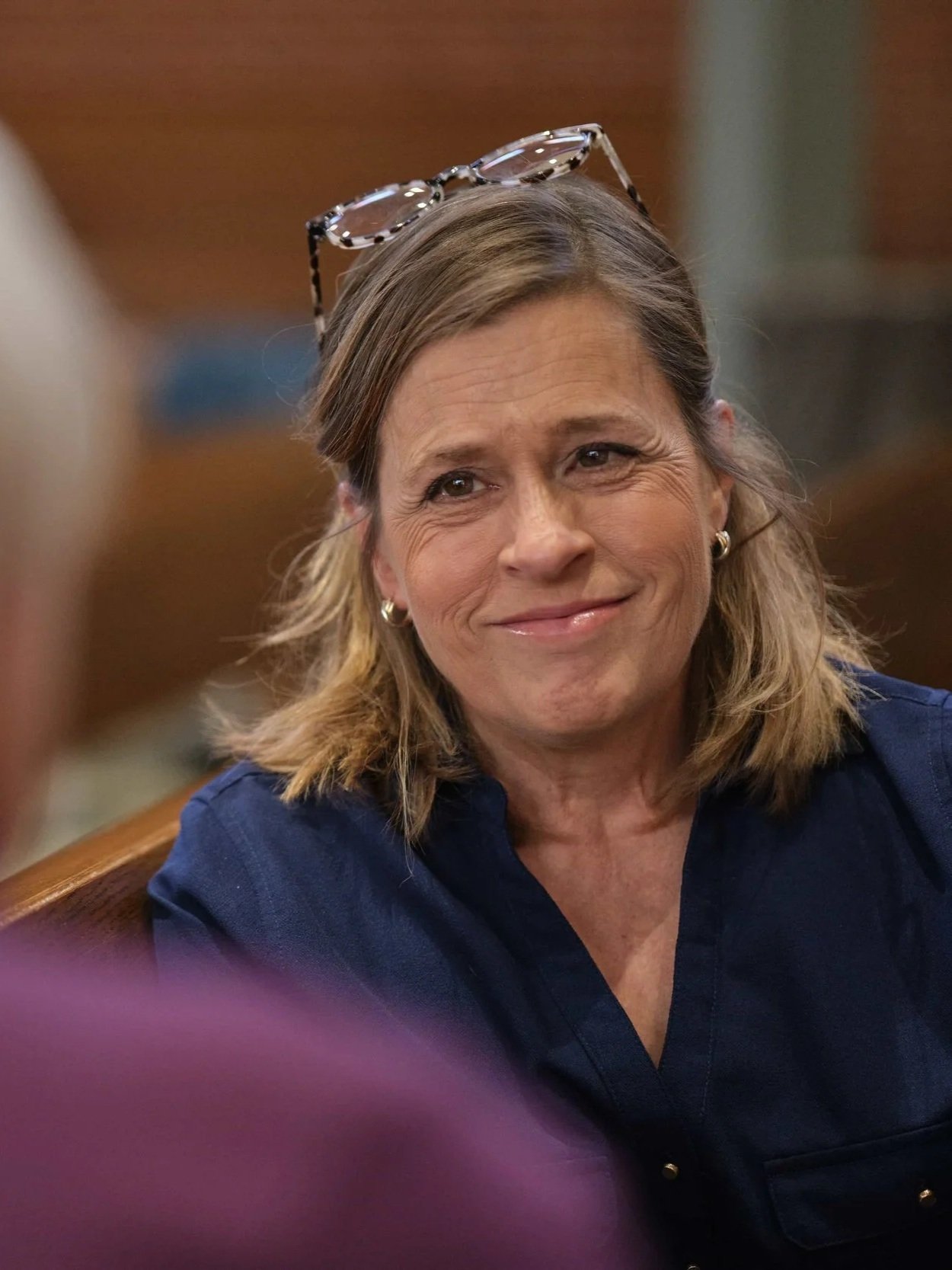 A member of Good Company, Ames Women's Choir with light skin, shoulder-length light brown hair, wearing glasses on her head, earrings, and a dark blue shirt, smiling.