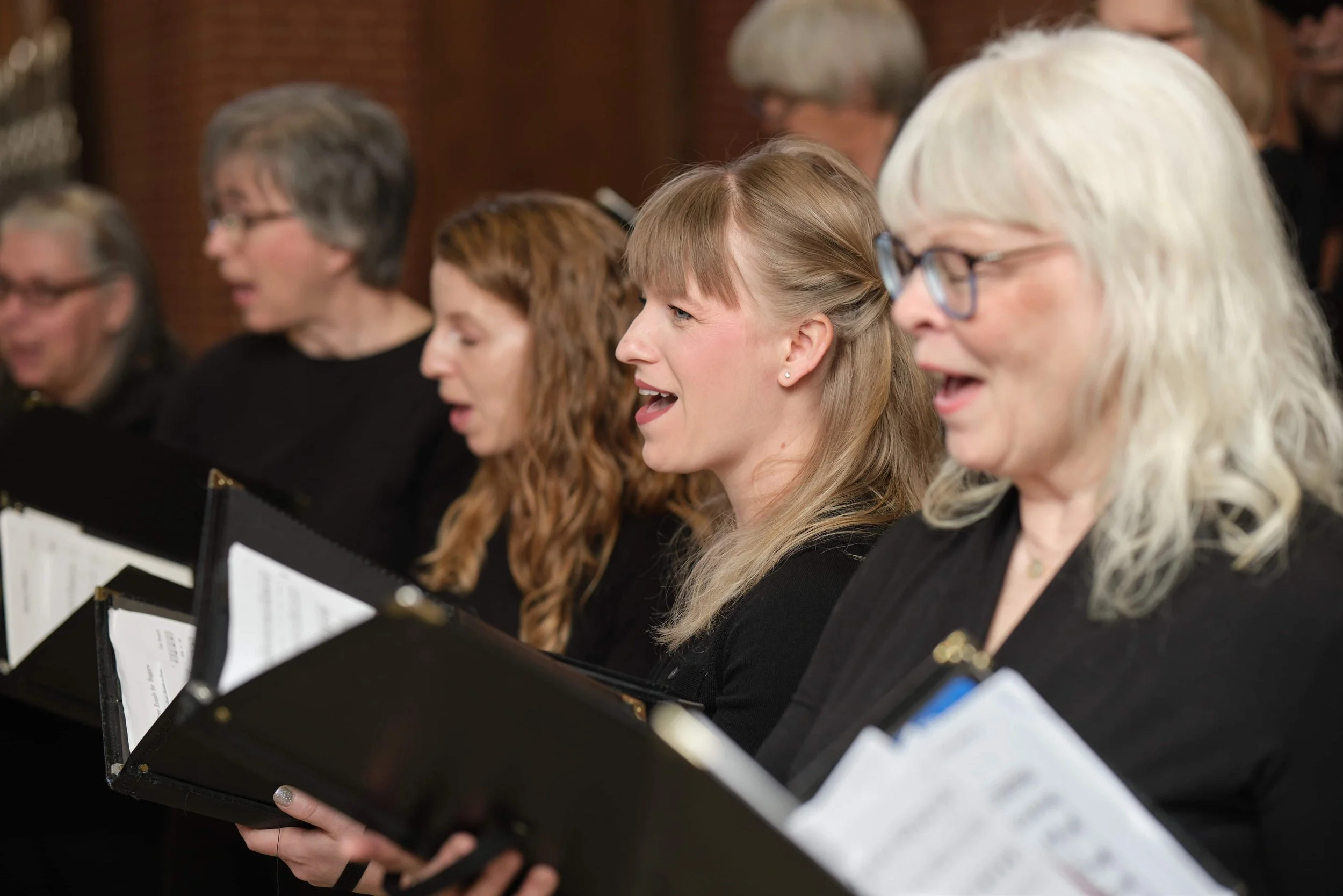 A group of women, all members of Good Company, Ames Women's Choir, singing from sheet music in a choir, sitting side by side in a formal setting.
