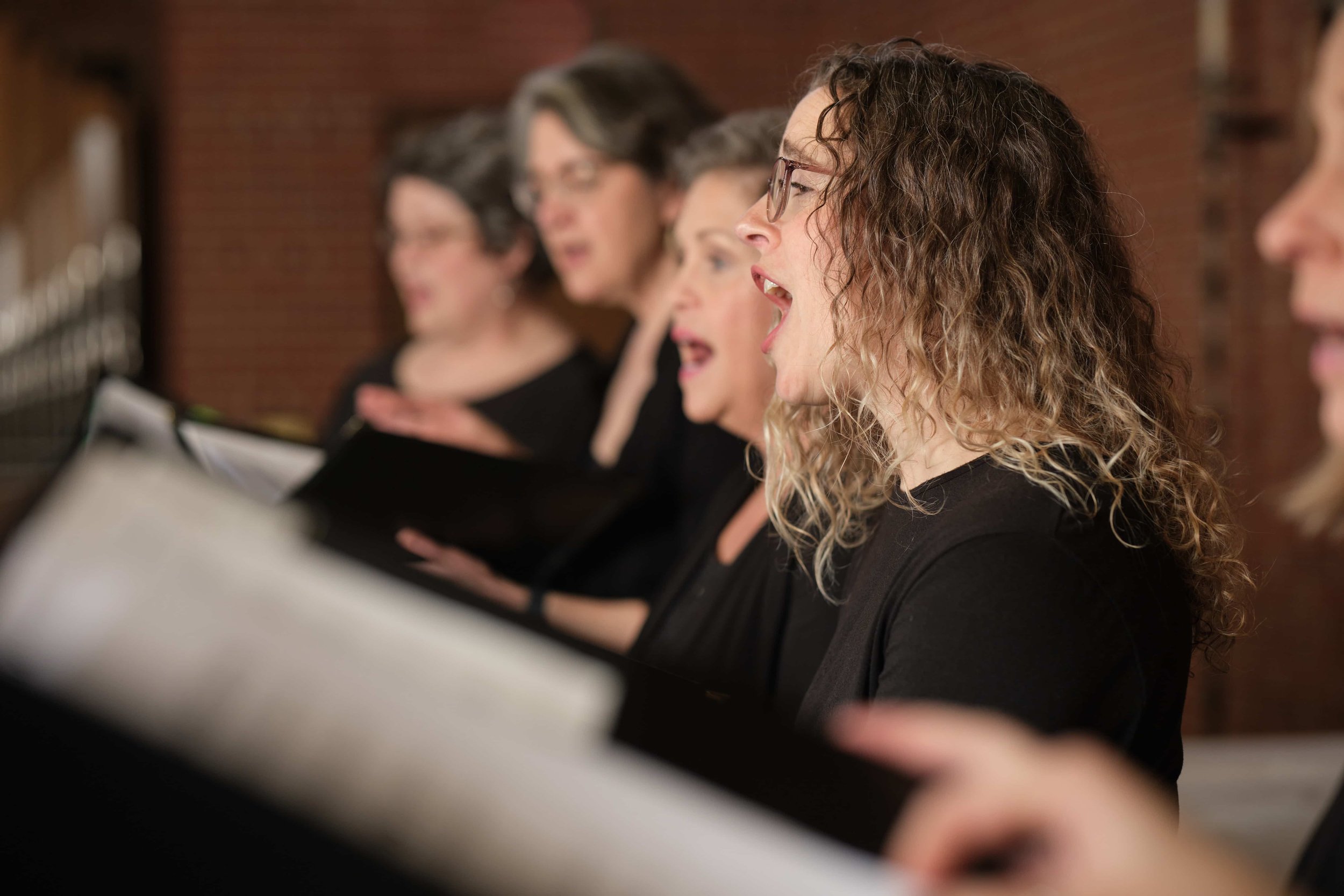 Side view of four women members of Good Company, Ames Women's Choir singing, with a brick wall in the background.