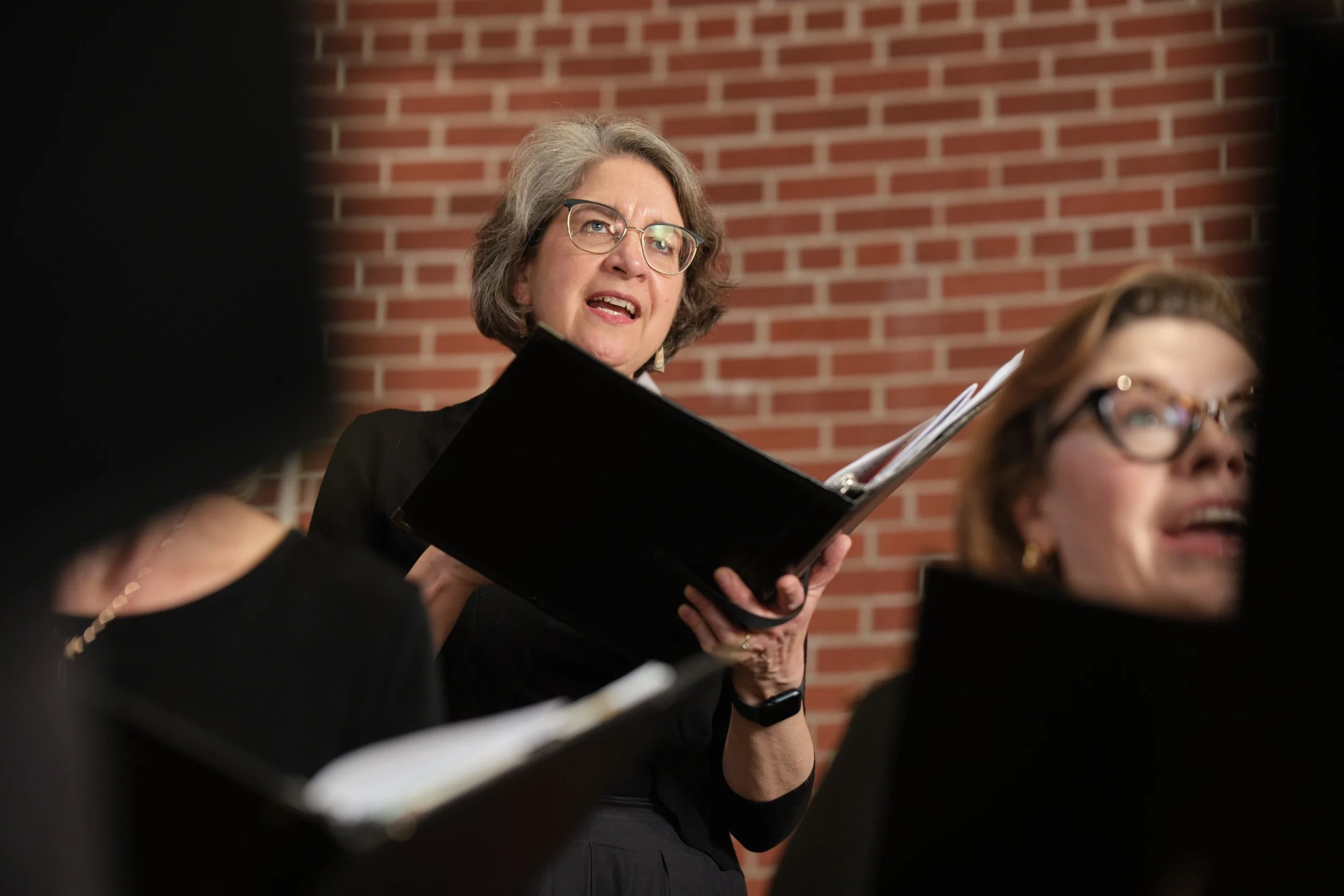A member of Good Company, Ames Women's Choir  with gray hair and glasses singing during a choir rehearsal, holding a black folder with sheet music, in front of a red brick wall.