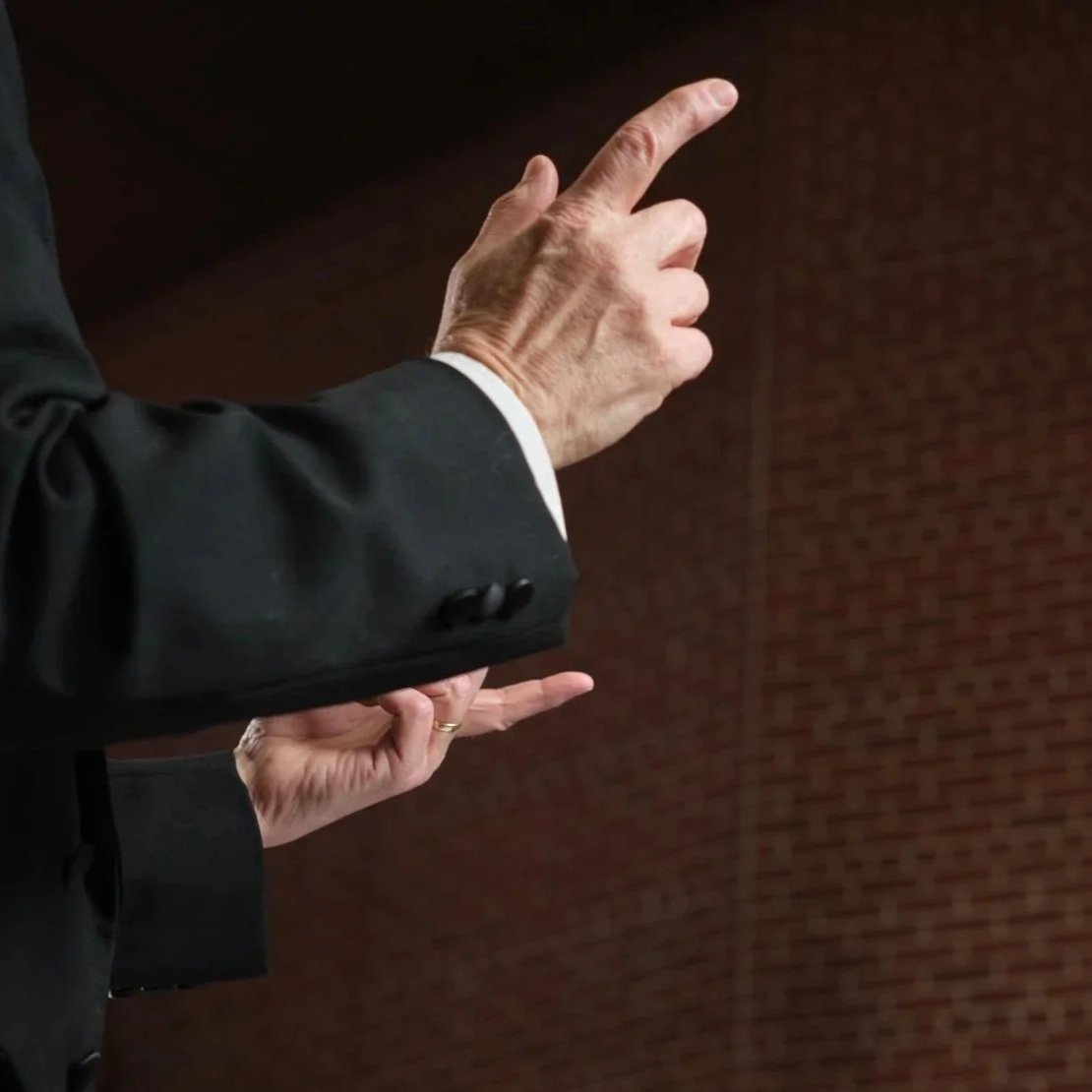 Close-up of Good Company director Steven Hoifeldt's hands making a gesture with the index finger pointing forward and the other fingers curled, dressed in a dark suit with a white shirt, against a brick wall background.