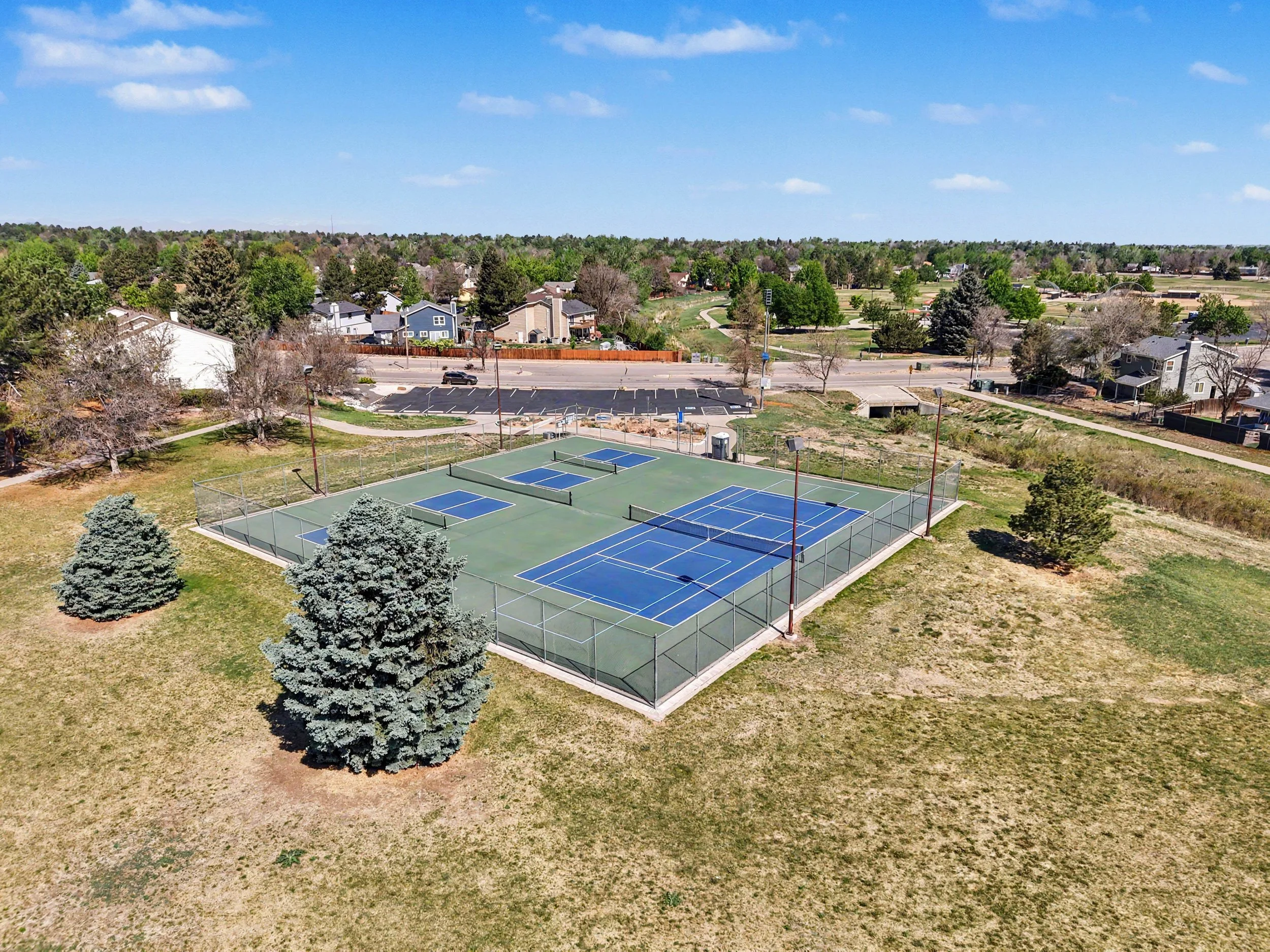 A park with tennis courts surrounded by grass and trees, located next to residential houses, under a partly cloudy blue sky.