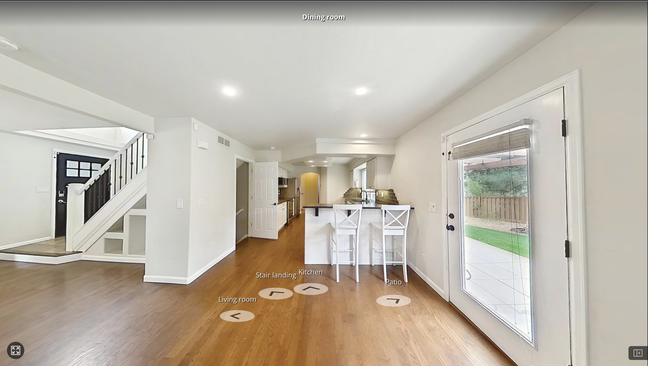 Interior view of a modern home with a living room, kitchen, patio, staircase landing, and dining room. Features include hardwood floors, white walls, large window with blinds, and a sliding glass door to the patio with a view of a backyard.