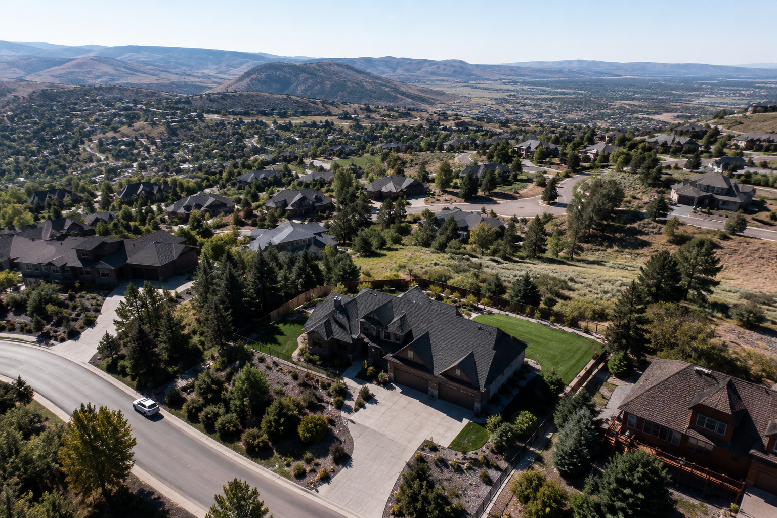 Aerial view of a suburban neighborhood with houses, trees, and a mountainous landscape in the background, showing a large house with a manicured lawn and driveway.