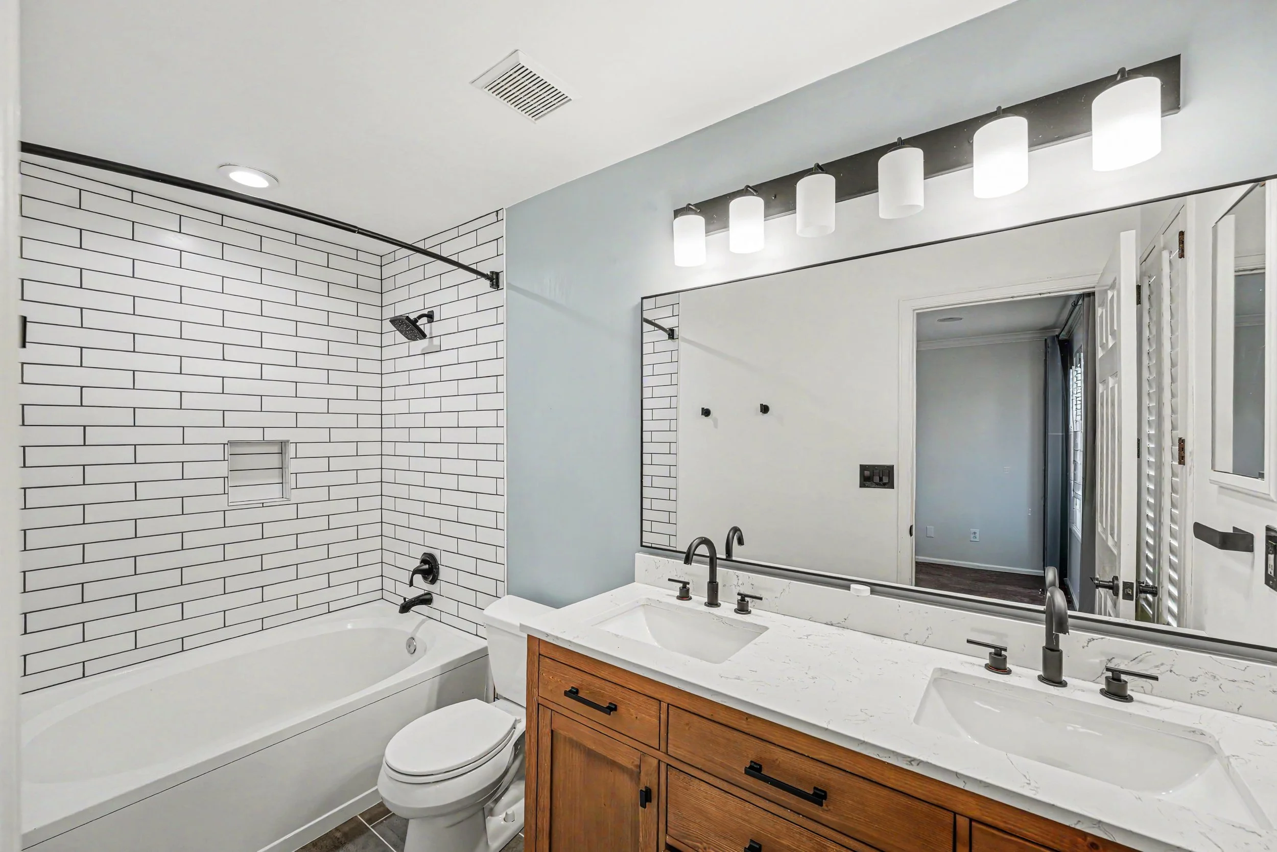 Bathroom with white subway tile shower area, black fixtures, large mirror, and wooden vanity with marble countertop.