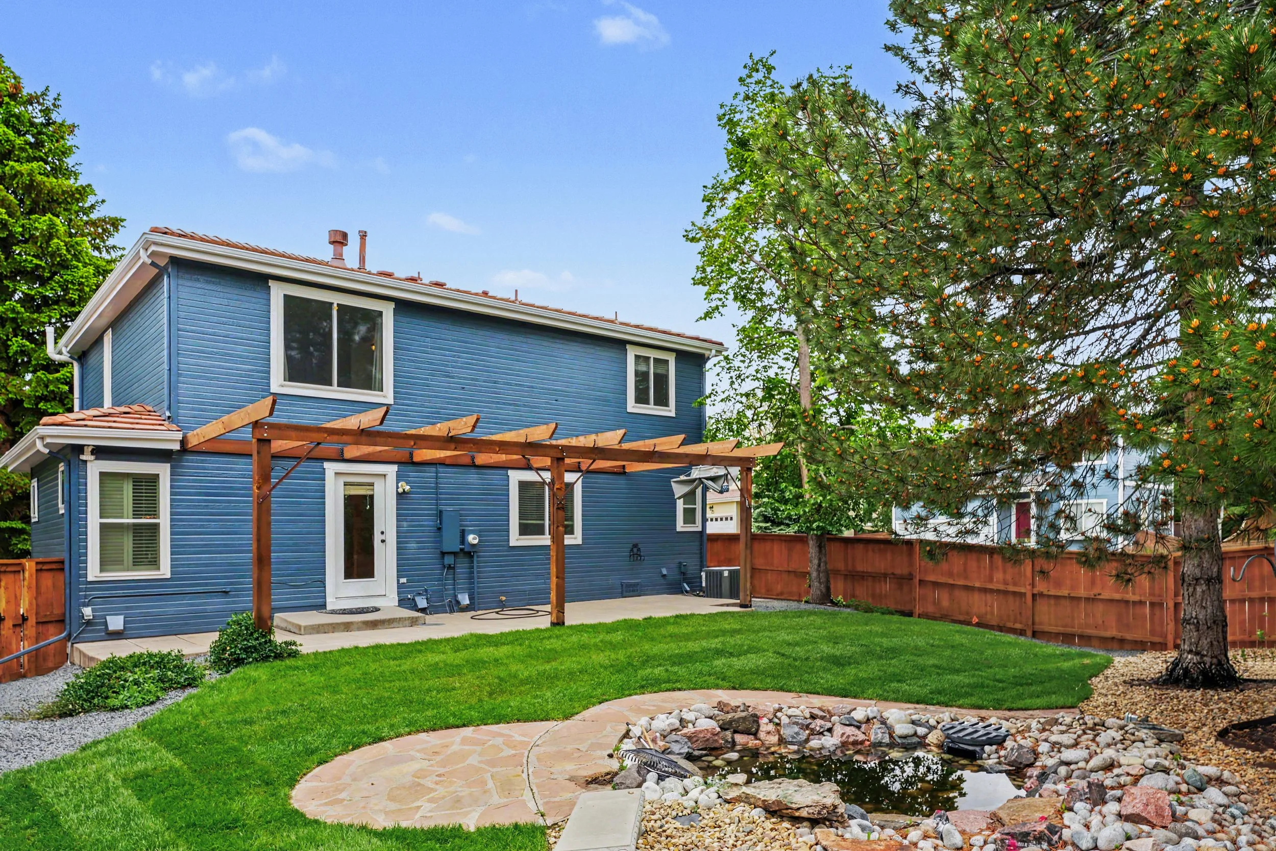 A backyard with green grass, a small pond surrounded by rocks, a concrete patio, and a blue two-story house with several windows. There is a wooden pergola frame near the house, a wooden fence, and mature trees, including one with small yellow cones or flowers. The sky is partly cloudy.