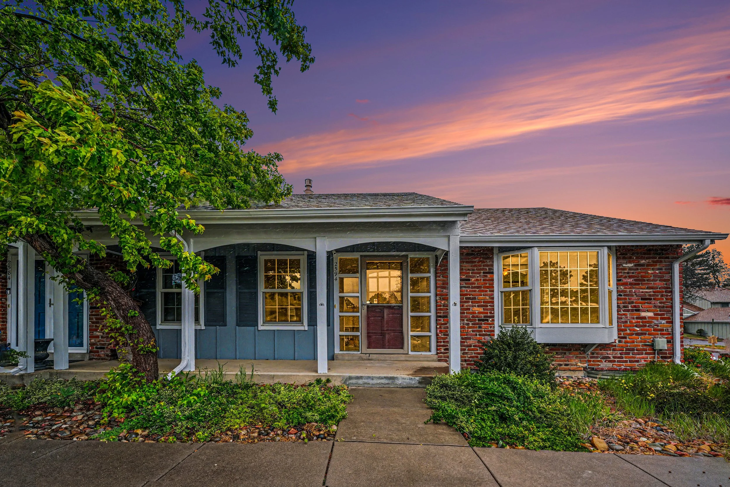 A house with a brick and vinyl exterior, large front windows, a wooden front door, and a porch. There is a tree with green leaves partially covering the left side of the house and a colorful sunset sky in the background.