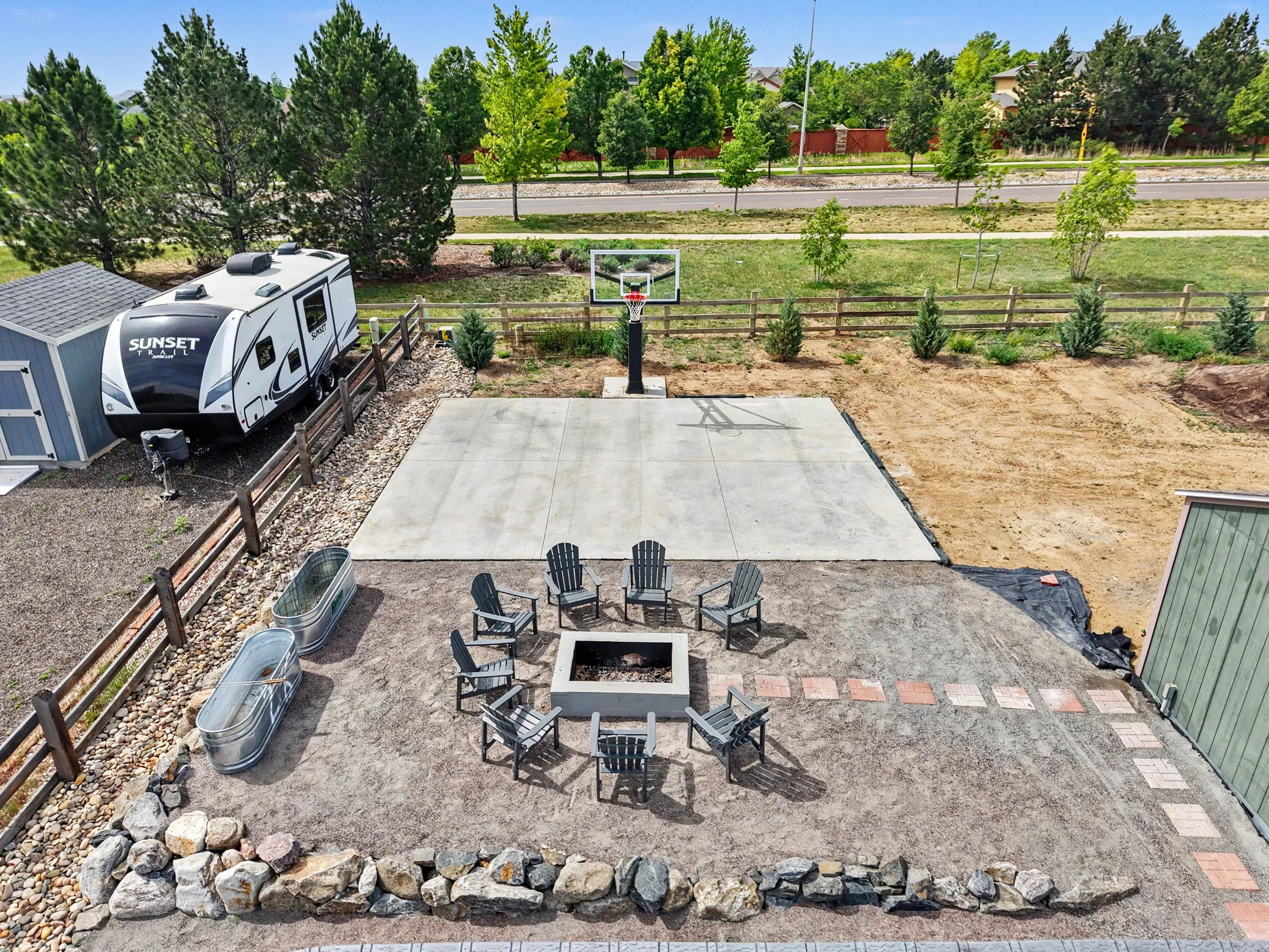 View of backyard with patio, outdoor seating, raised fire pit, trees, and a basketball hoop, with a backyard fence and street in the background.