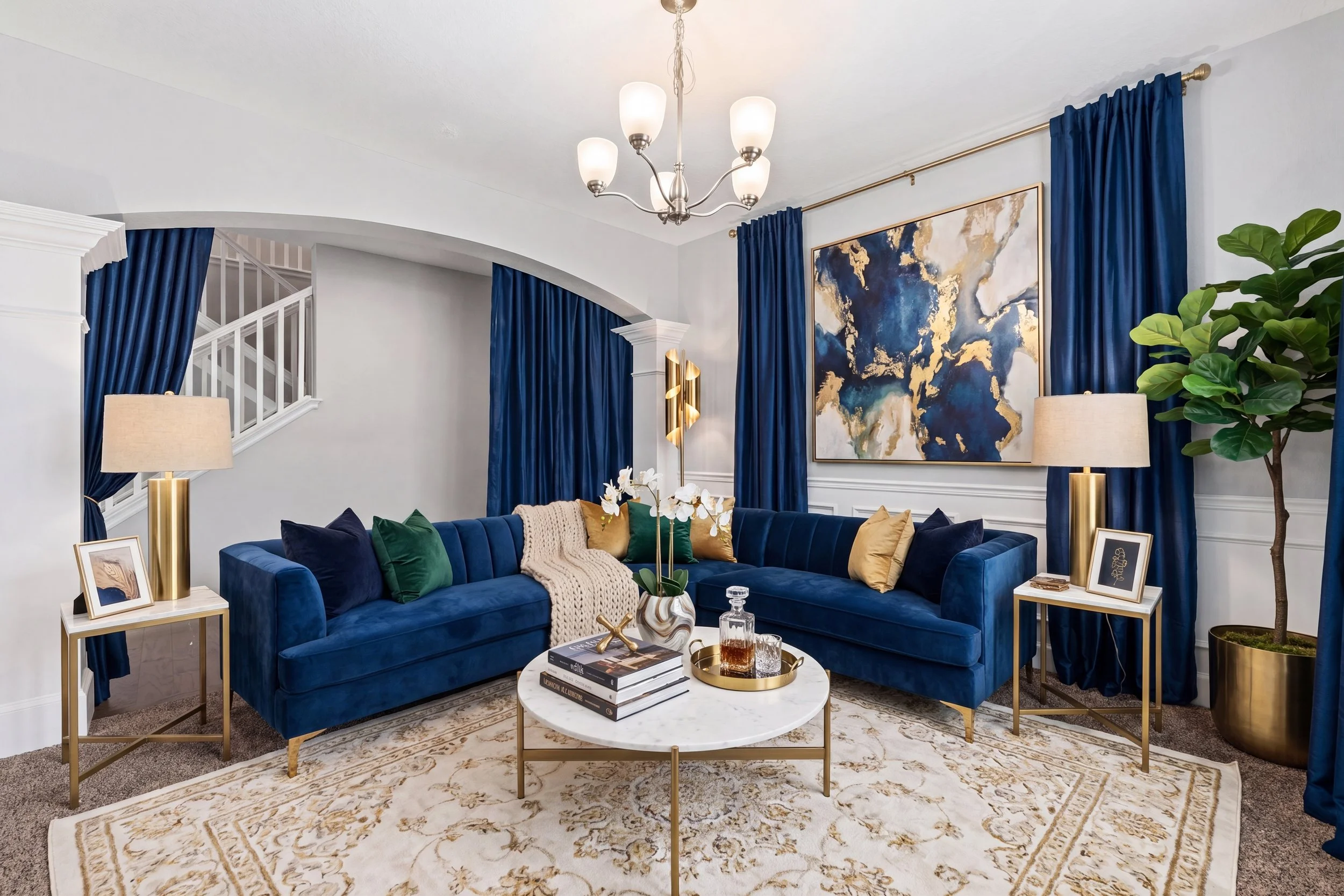 Living room with blue velvet sofa, gold accent tables, abstract artwork, blue curtains, potted plant, modern lighting, and marble coffee table.