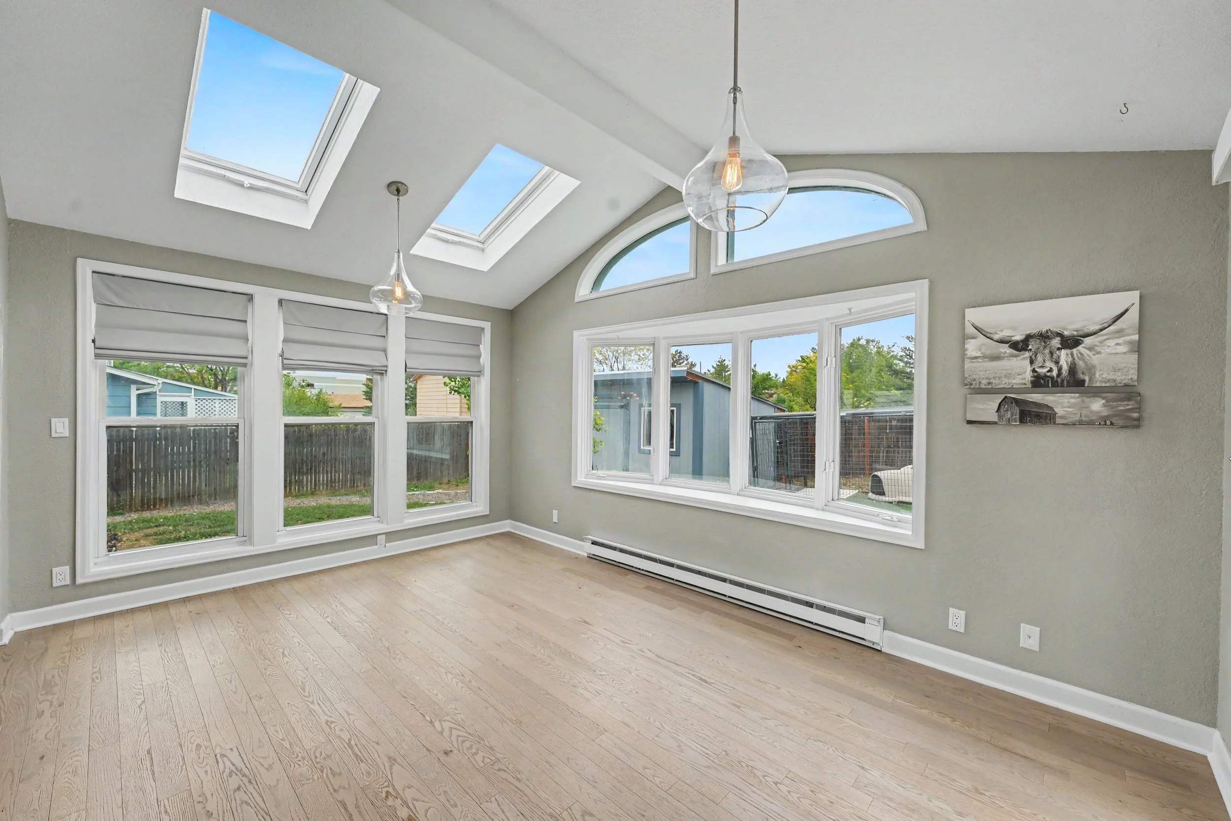 Bright living room with large windows, skylights, and a ceiling light fixture, featuring light wood flooring and neutral-colored walls.