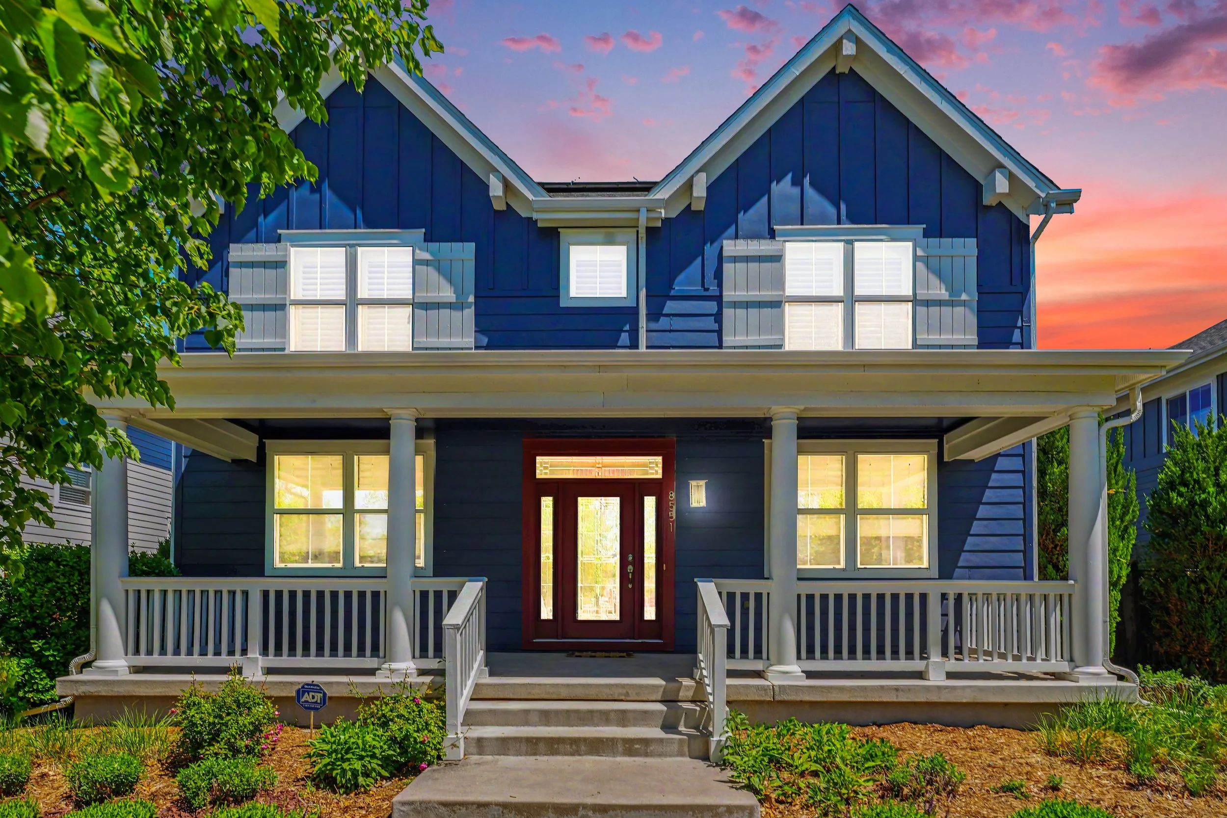 Blue two-story house with a front porch, white columns, and railing, set against a sunset sky, with windows and a red front door surrounded by greenery and landscaping.