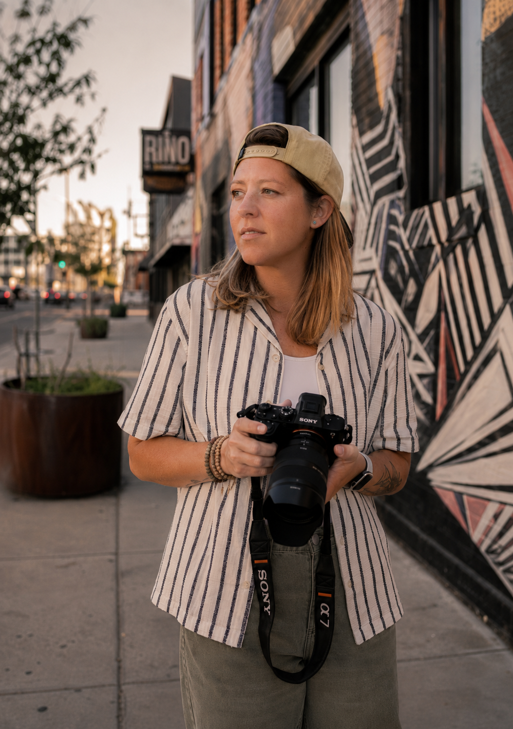 A woman with shoulder-length brown hair wearing a beige and black striped shirt, khaki pants, and a backward beige baseball cap, holding a photographer's camera, standing on a sidewalk in front of a mural.