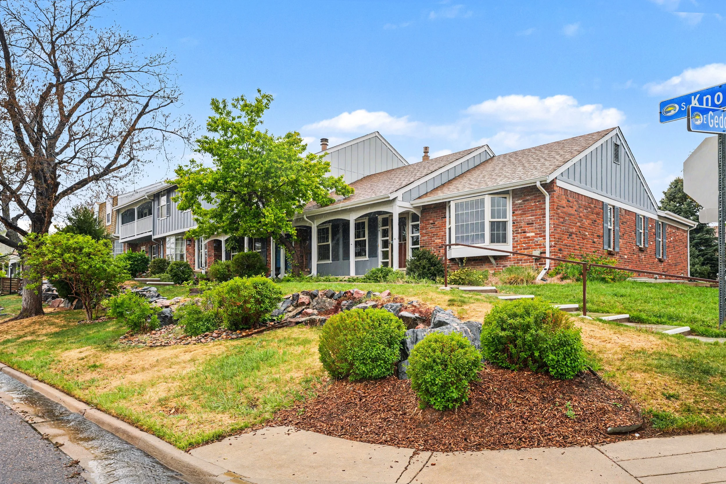 A residential neighborhood with brick and gray houses, a small front yard with bushes, trees, and a sidewalk, along with a blue street sign on the corner of the street.