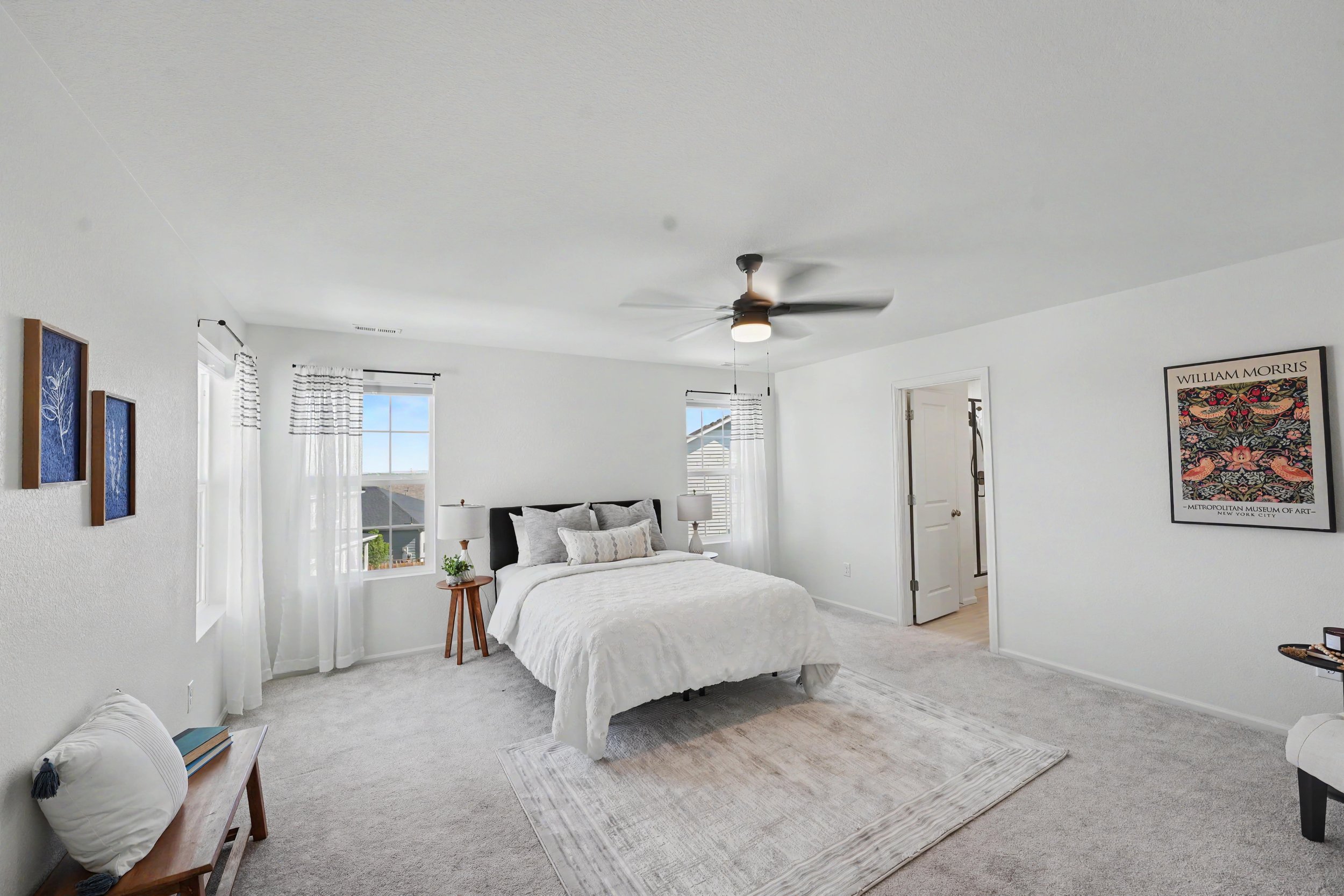 Bright bedroom with white walls, ceiling fan, large window with striped curtains, bed with white bedding, bedside tables with lamps, wall art, and a bench with a pillow at the foot of the bed.