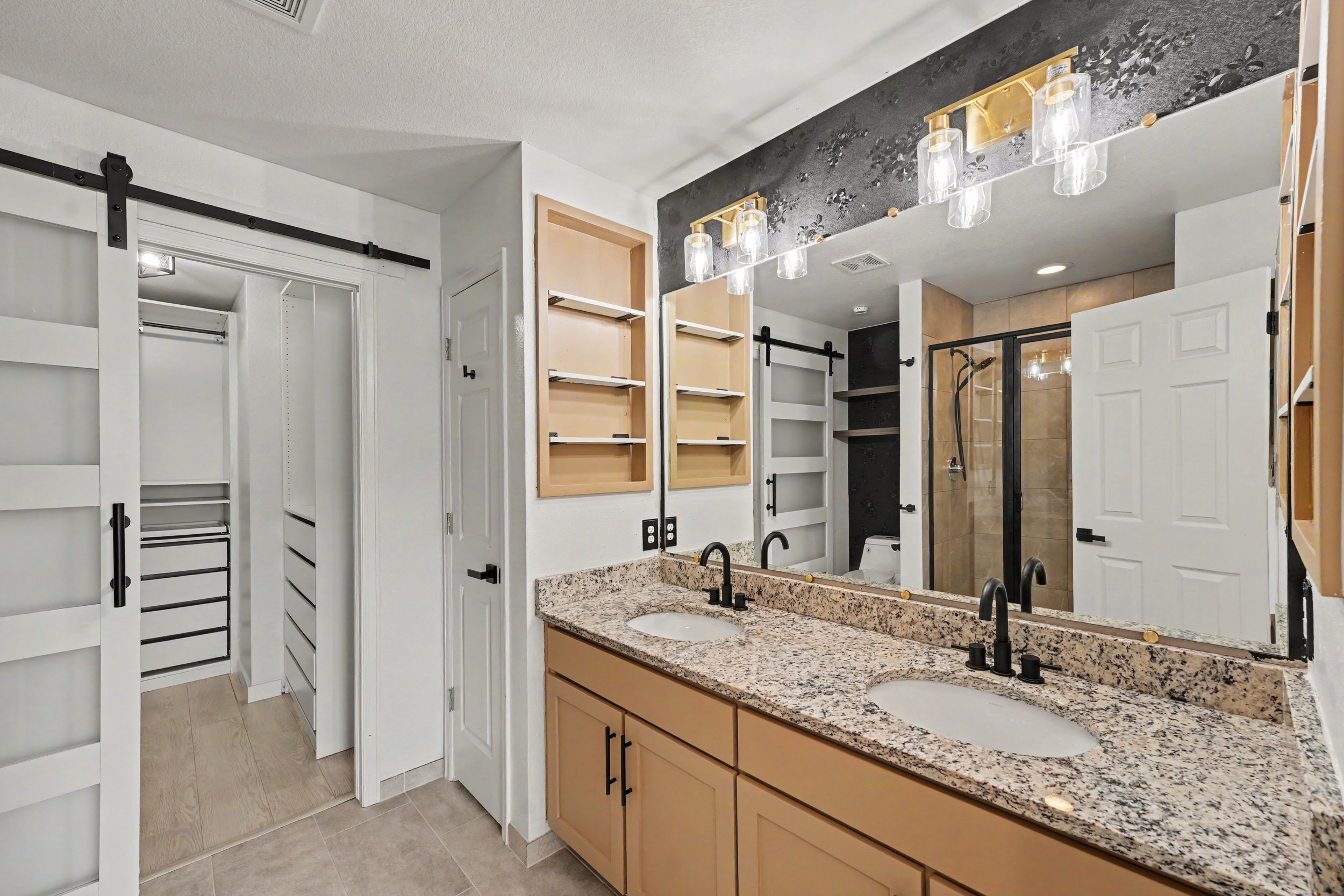 Bathroom with granite countertop, large mirror, black fixtures, and a walk-in shower with a glass door. There are open and closed closet doors, some shelving, and black sliding door hardware.
