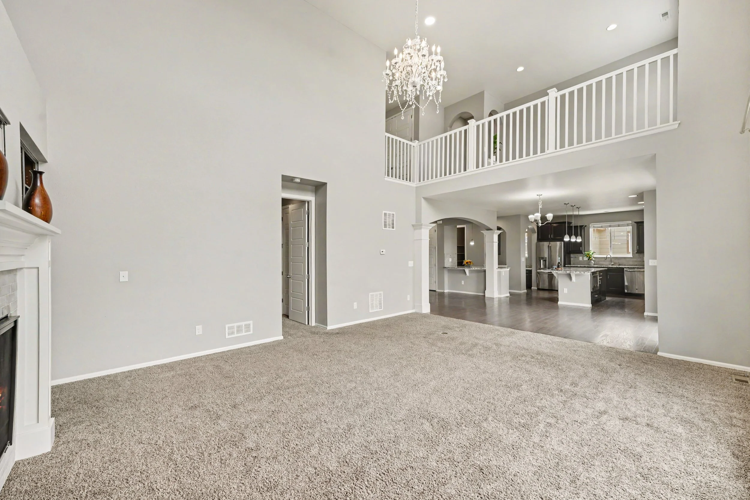 Interior view of a spacious living room with beige carpet, white walls, a white fireplace mantel with decorative vases, and a staircase leading to an upper level. The open kitchen and dining area are visible in the background, with dark cabinets, stainless steel appliances, and pendant lighting.