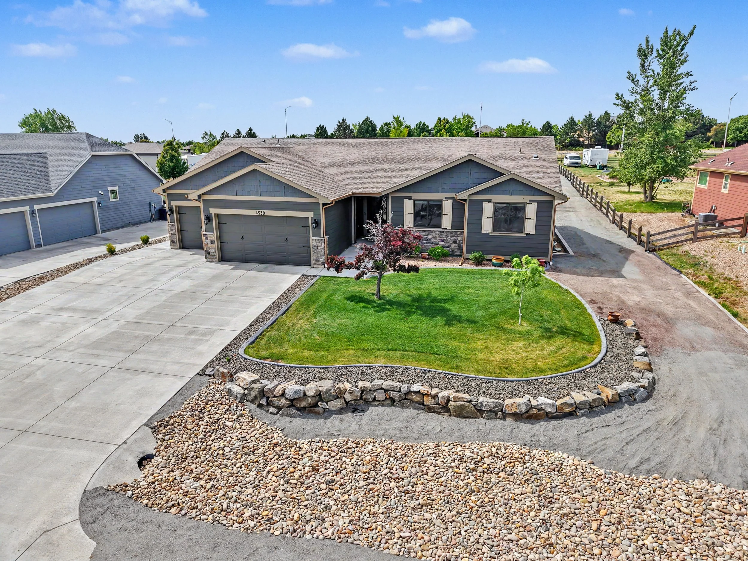 A residential house with a gray exterior, stone accents, and a motorcycle parked beside it; a driveway with a small landscaped area featuring a tree, shrubs, and decorative rocks; neighboring houses and greenery in the background under a blue sky with clouds.
