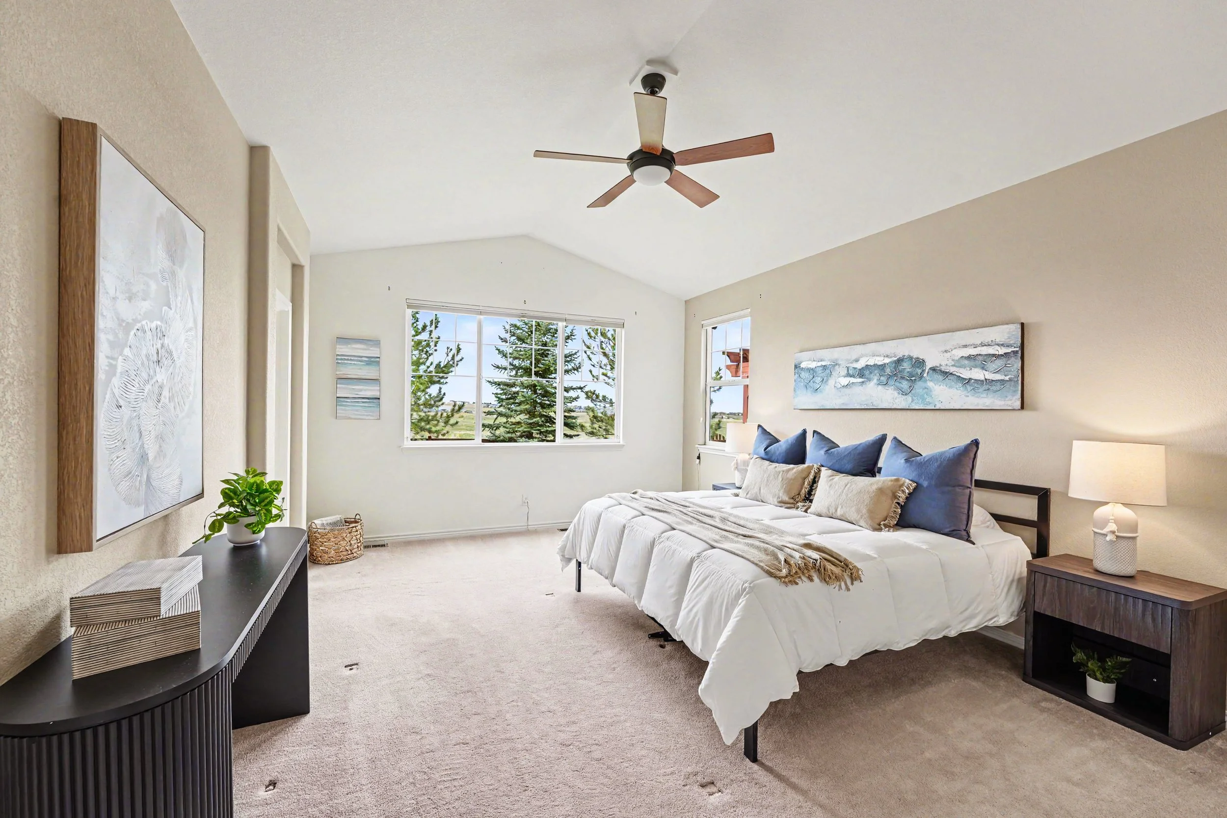 A bedroom with a white bed, blue pillows, abstract wall art, a black console table with a plant, and a ceiling fan with wooden blades. Large windows show trees outside.