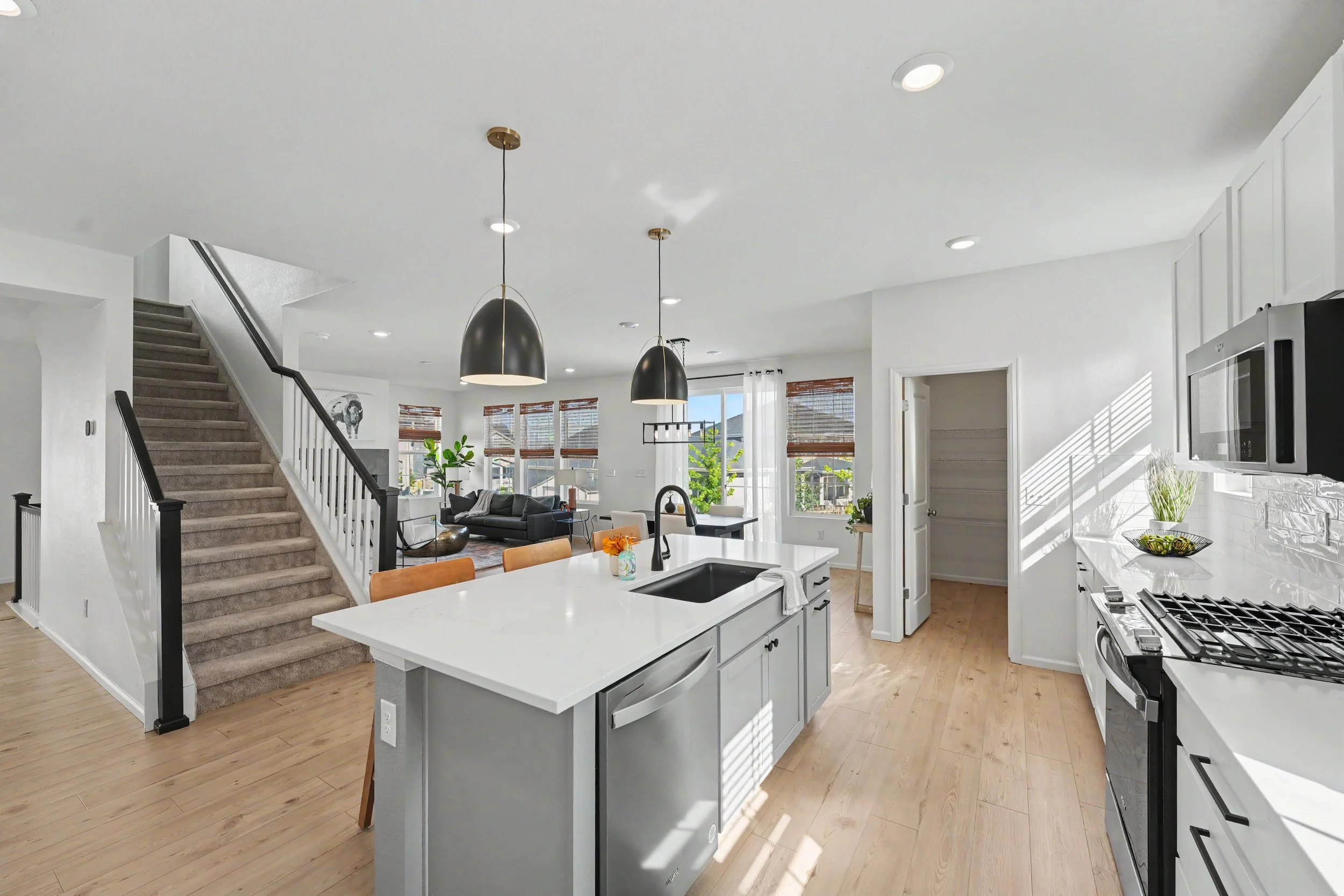 Open-concept living room and kitchen with white cabinets, a kitchen island, and black pendant lights, with large windows and staircase.