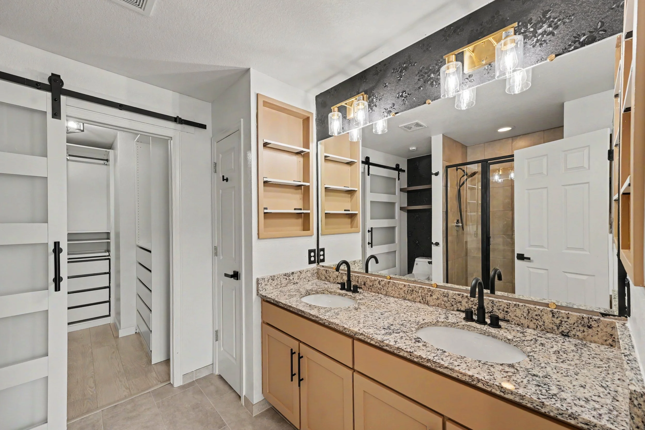 Bathroom with granite countertop, double sinks, black fixtures, wooden cabinets, large mirror, and a walk-in shower enclosed in glass.