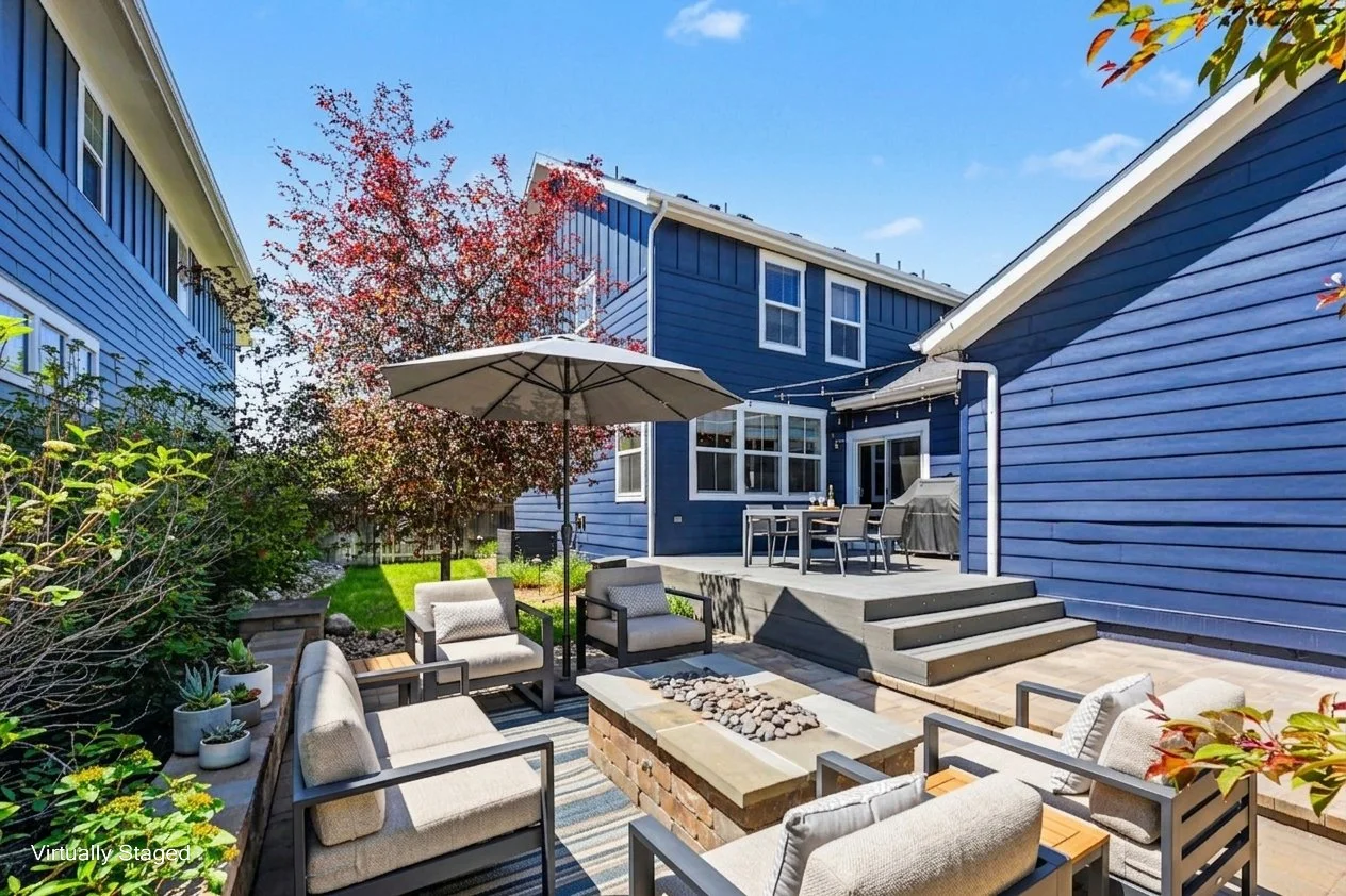 Backyard patio with outdoor seating, a fire pit, an umbrella, a dining table with chairs, a barbecue grill, and a blue house with a deck and string lights, on a sunny day with a tree showing fall colors.