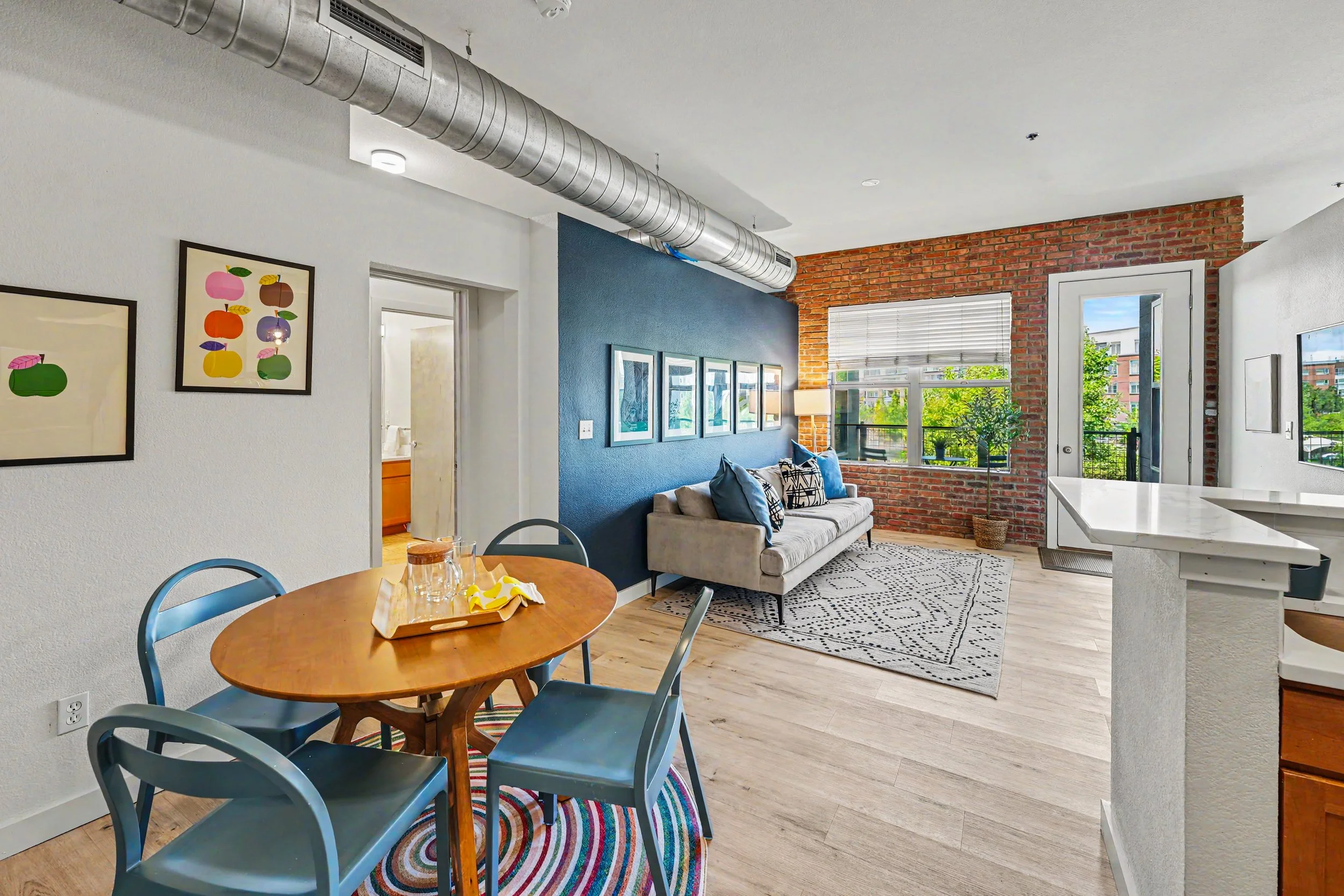 Open-concept living room and dining area with brick wall, large windows, and modern furniture.