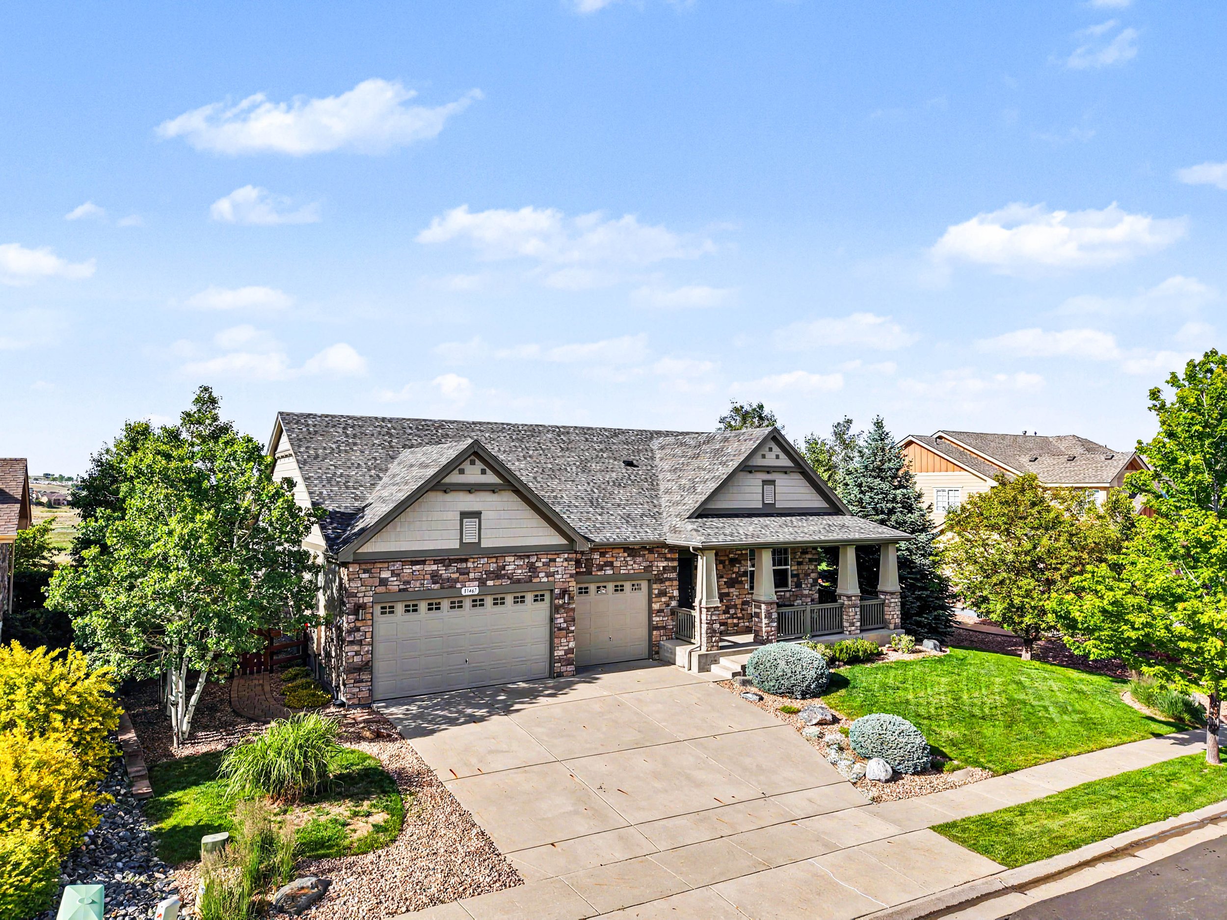 A suburban house with a gray shingle roof, a two-car garage, and a front porch. The front yard has trees, shrubs, and a well-maintained lawn, with a concrete driveway extending to the sidewalk. The sky is clear with some clouds.