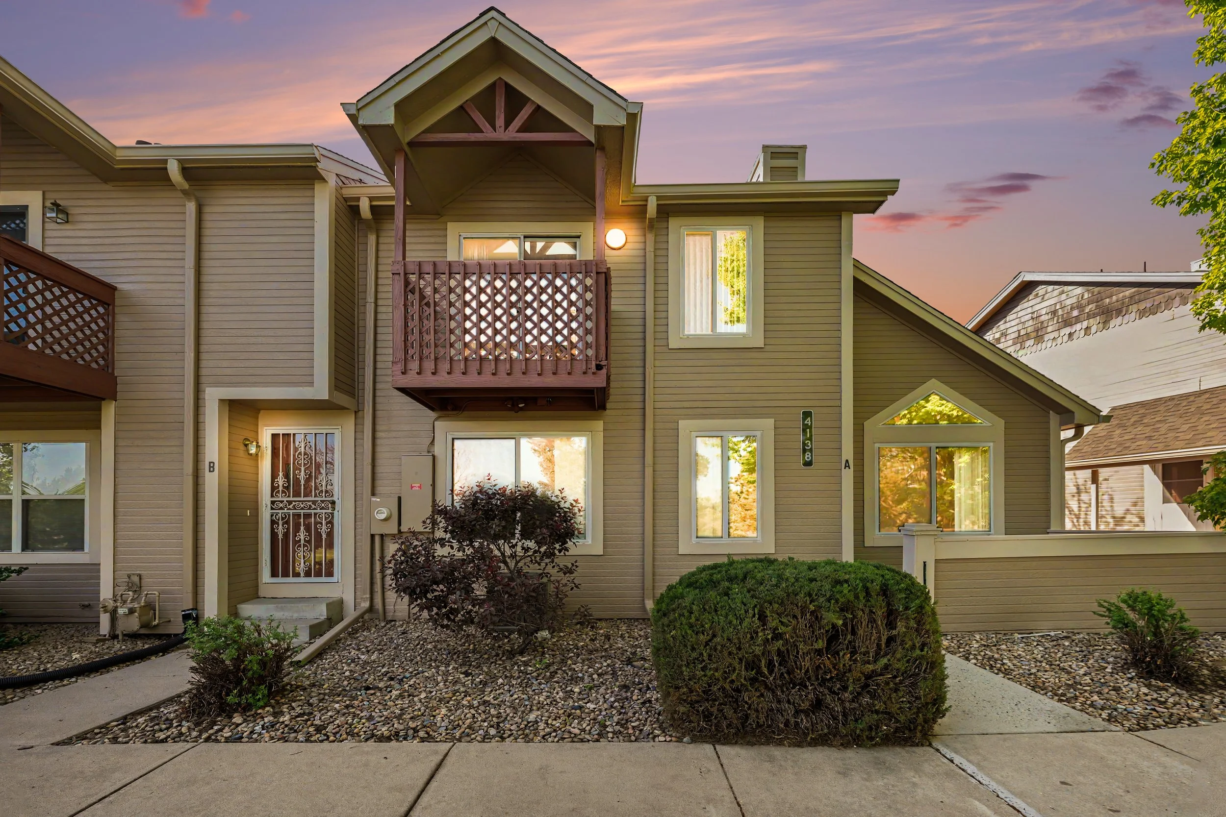 Rear view of a multi-story beige apartment building with small balconies and a variety of window styles, surrounded by landscaped bushes, trees, and a gravel area, during sunset with a colorful sky.