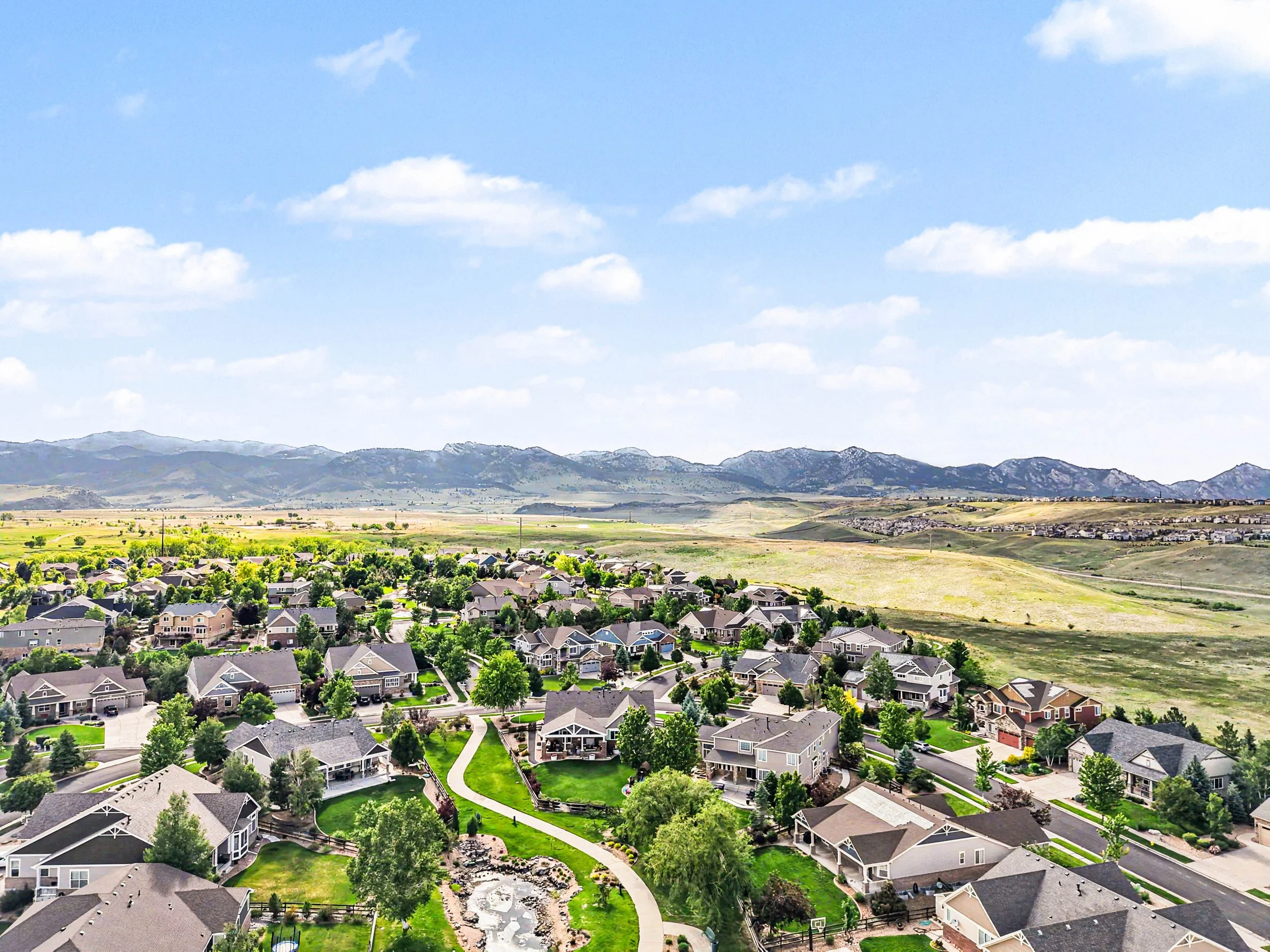 Aerial view of a suburban neighborhood with houses, trees, a backyard playground, and a green lawn. In the background, there are distant mountains under a blue sky with scattered clouds.