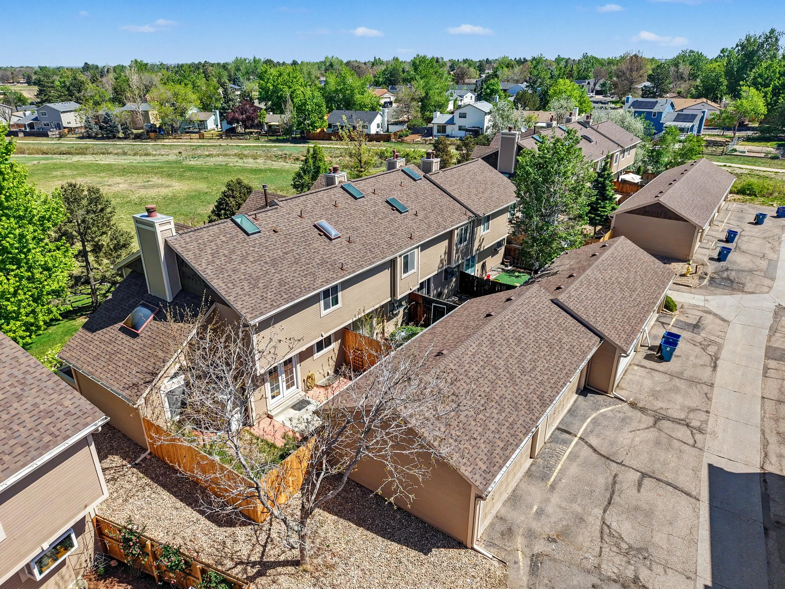 A residential neighborhood with houses and townhomes with brown shingle roofs, green trees, a grassy field, and a parking lot with blue trash cans, under a clear blue sky.