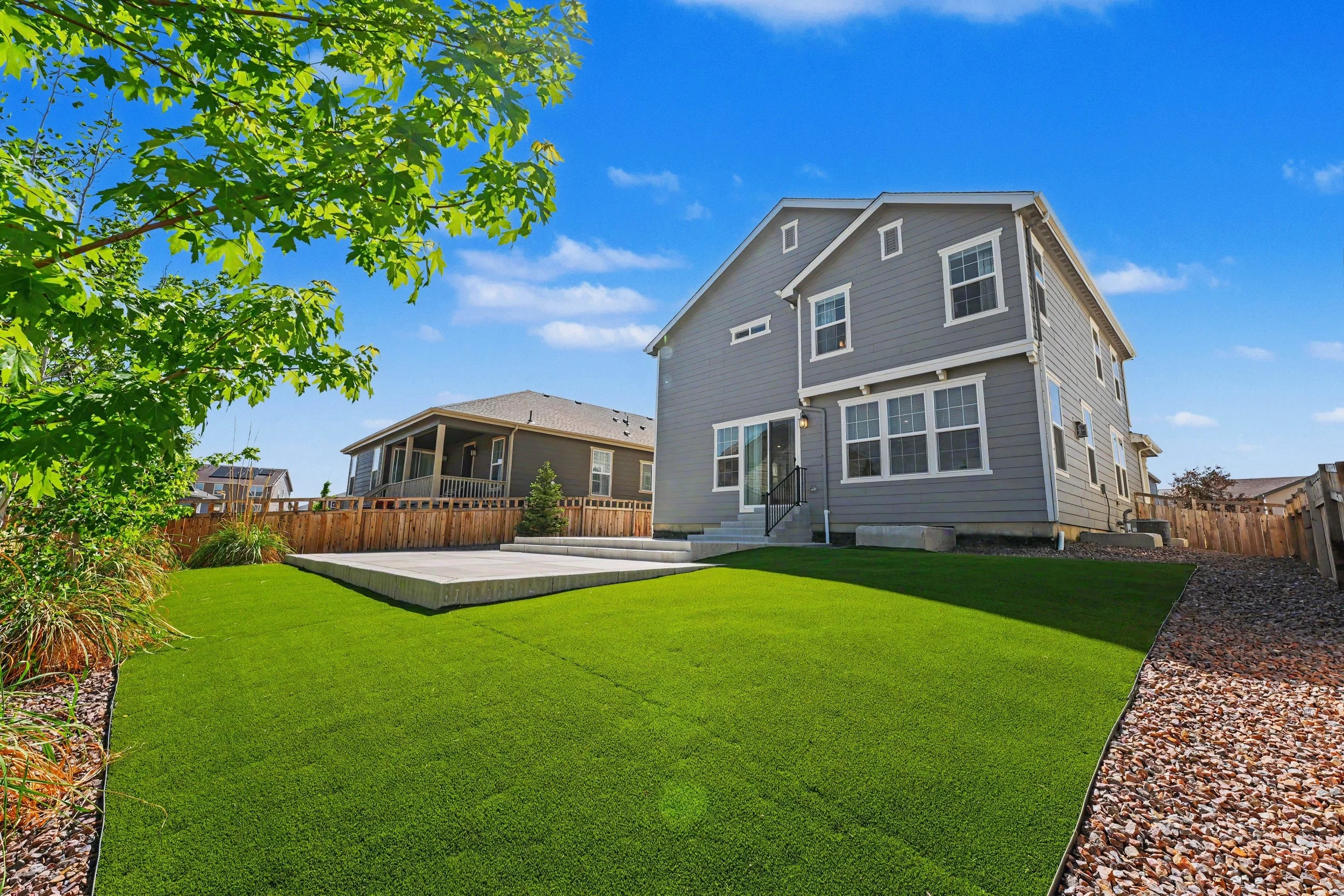 A backyard with a neatly manicured green lawn, a wooden deck, a gray house with multiple windows, and a wooden fence. A tree with green leaves is visible on the left side, and the sky is blue with a few wispy clouds.