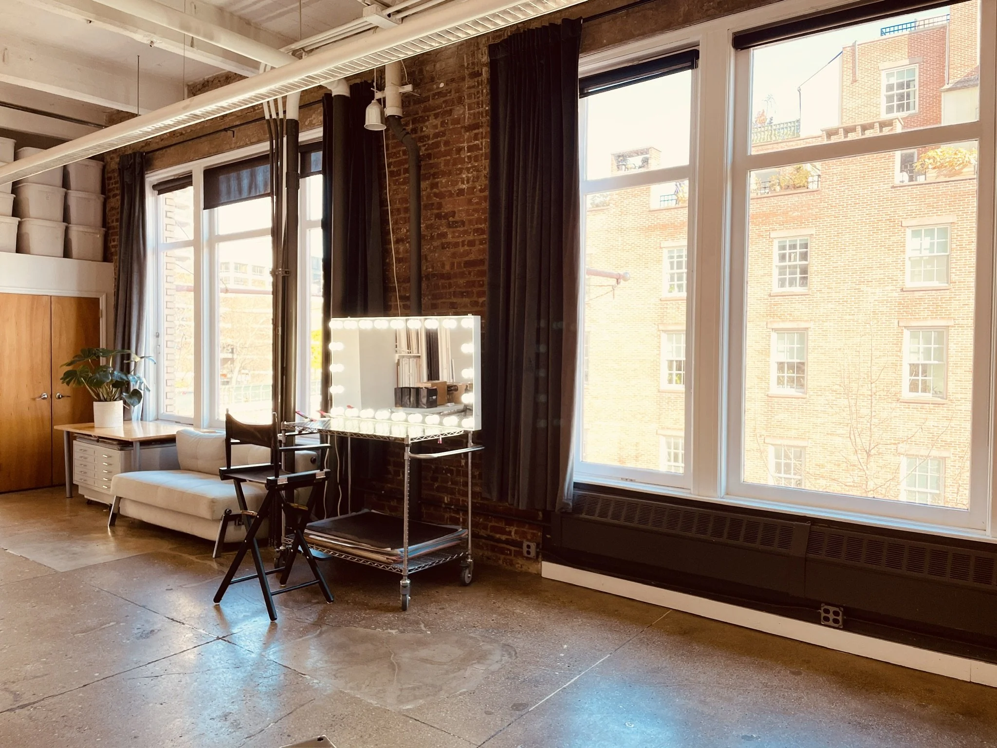 Interior of a loft-style room with large windows, brick walls, black curtains, a white couch, a potted plant, and a vanity mirror with lights.