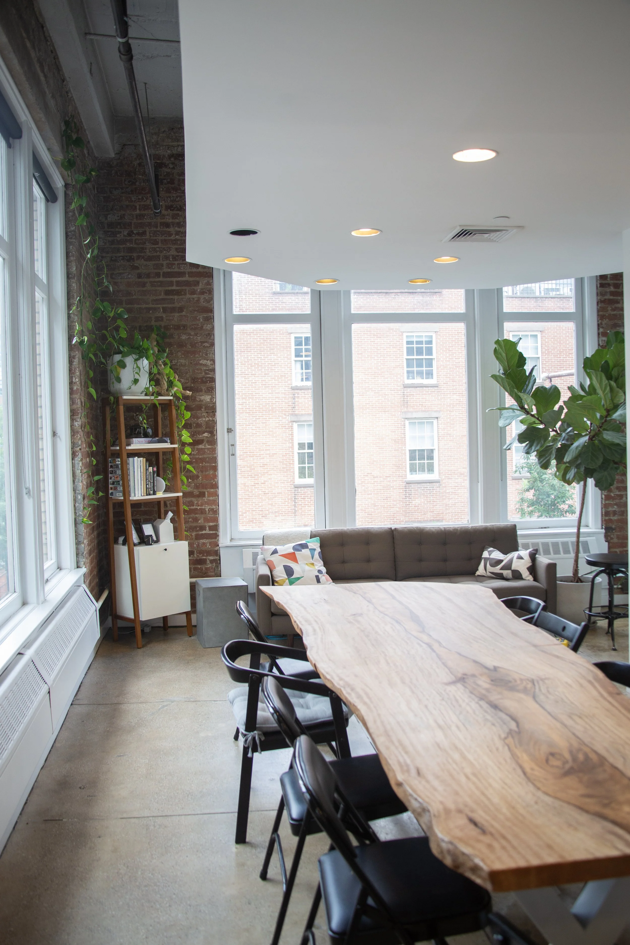 A meeting room with a large, natural wood table, black chairs, a gray couch with colorful pillows, large windows, exposed brick wall, indoor plants, and ceiling lights.